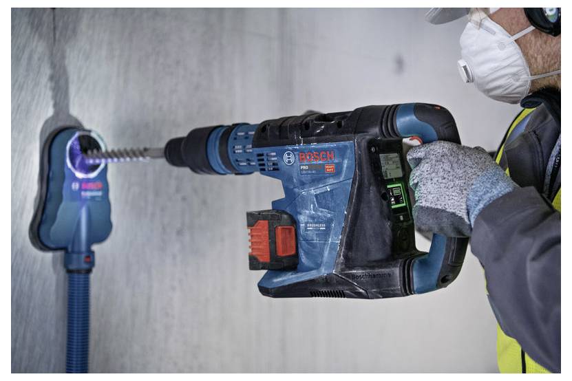 Un operaio sta praticando un foro in una parete di calcestruzzo con un trapano elettrico Bosch mentre indossa guanti, una mascherina e dispositivi di protezione individuale.