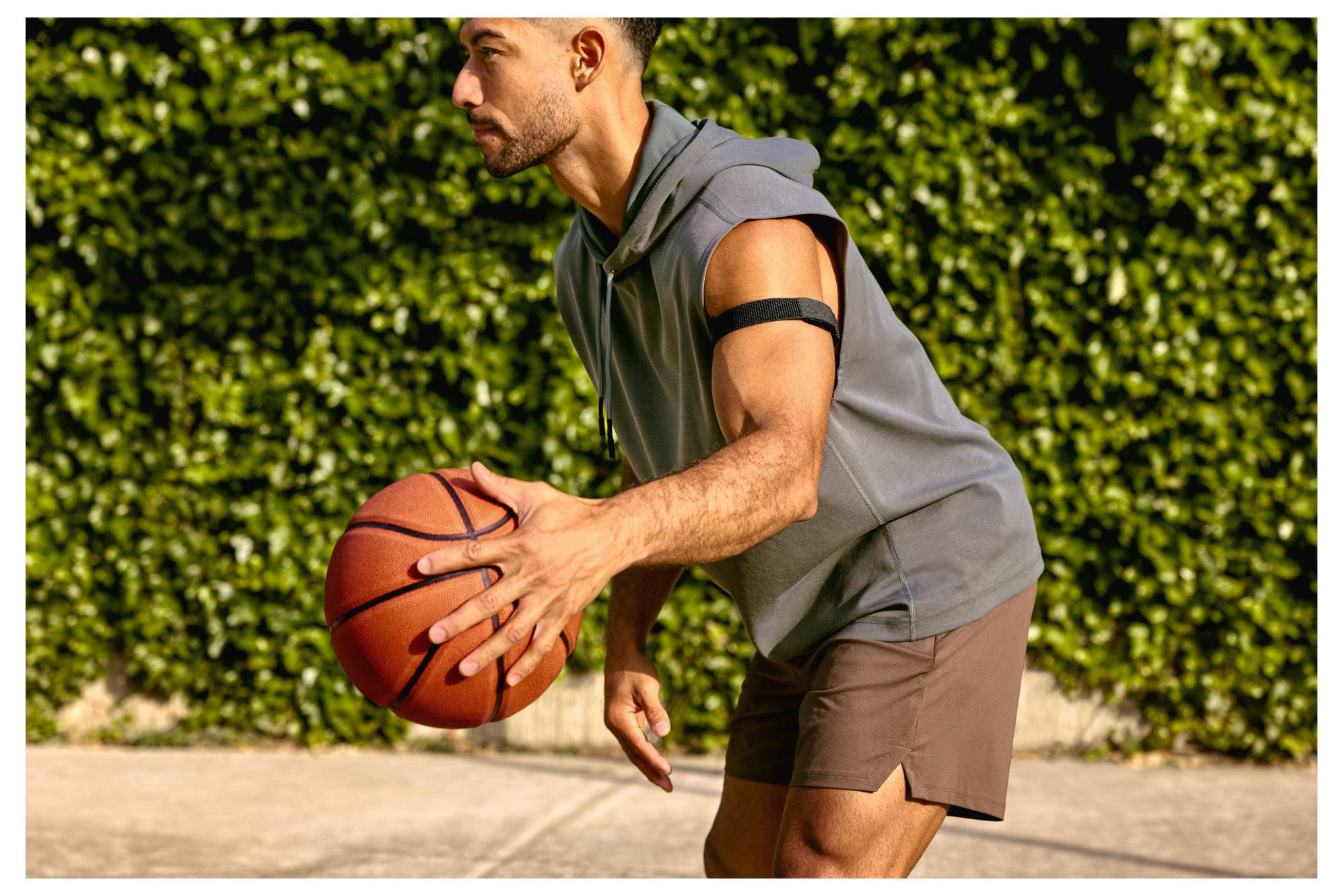 Uomo che palleggia un pallone da basket all'aperto su un campo illuminato dal sole, indossando una felpa senza maniche e pantaloncini, in posizione di attesa.