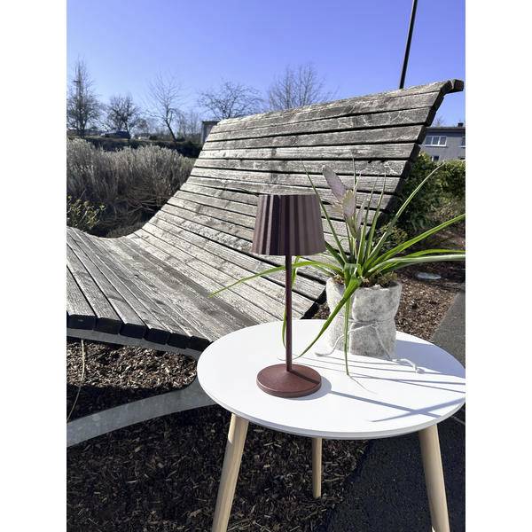 Area esterna con sedute dotata di una panchina in legno curva dietro un piccolo tavolo bianco su cui sono appoggiati una lampada e una pianta in vaso sotto un cielo sereno.
