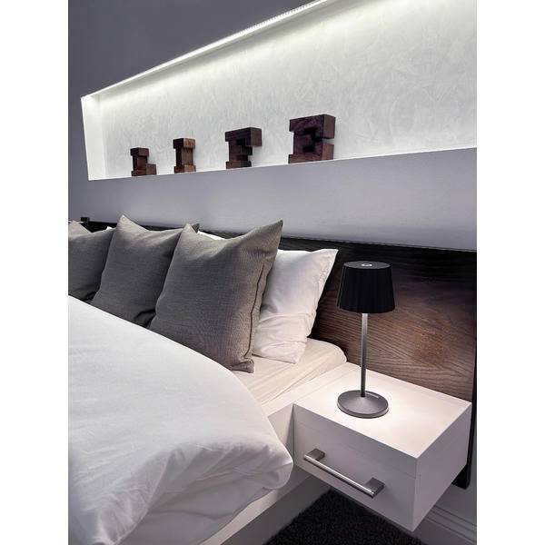 Camera da letto moderna con un letto perfettamente fatto, cuscini grigi, biancheria bianca e un comodino con una lampada nera sotto uno scaffale di testata illuminato.