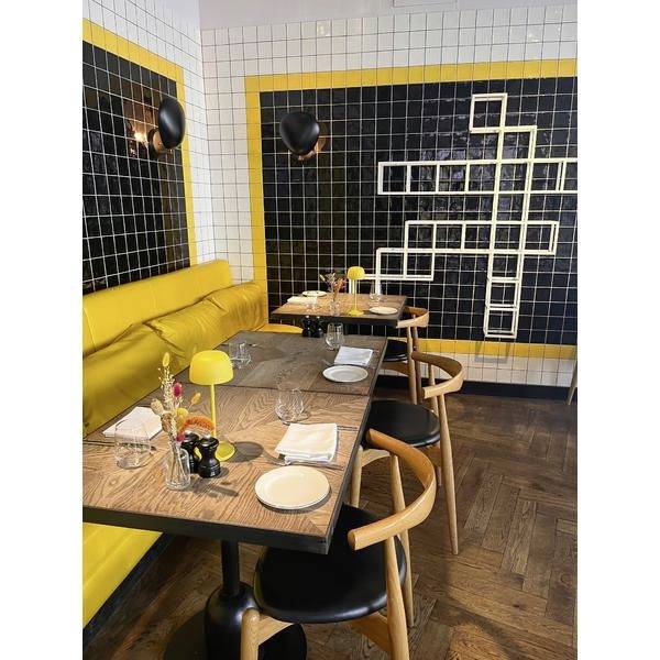 Angolo di ristorante moderno con sedute a panchina gialla, tavoli in legno apparecchiati per il servizio, e una parete con piastrelle nere caratterizzata da un design geometrico di quadrati bianchi.