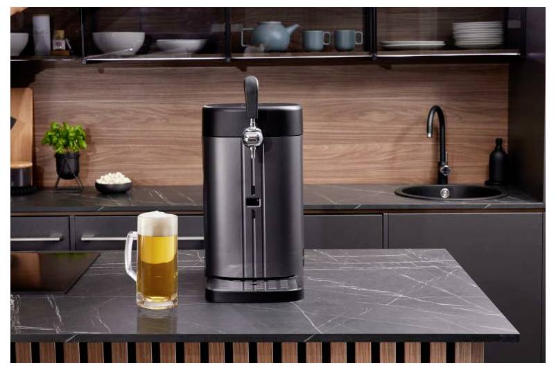 Dispenser di birra nero per la casa su un bancone della cucina accanto a una tazza di birra piena, con un lavandino e ripiani sullo sfondo.