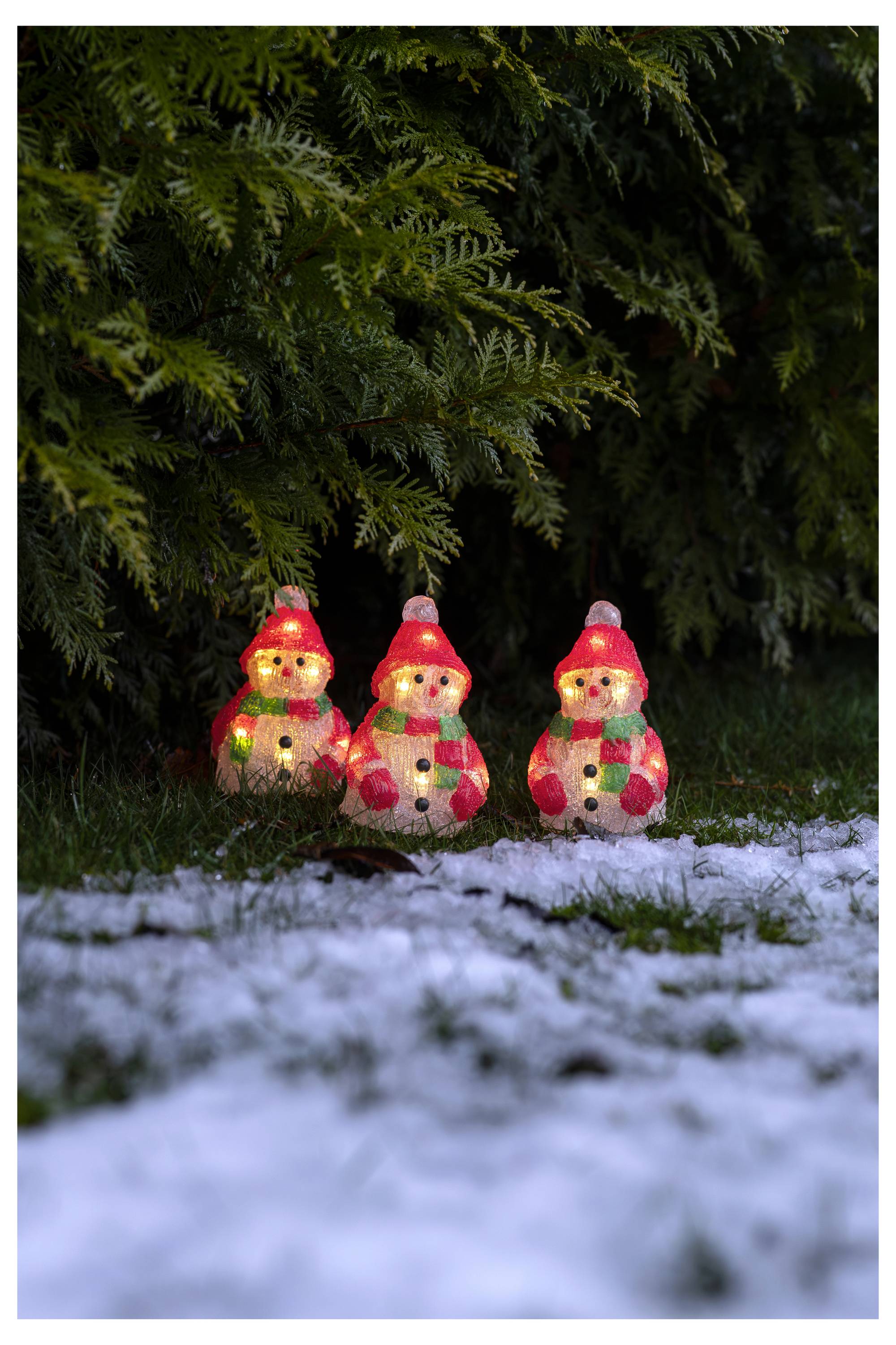 Tre pupazzi di neve luminosi con luci natalizie sono seduti sull'erba innevata sotto i rami scuri degli abeti.