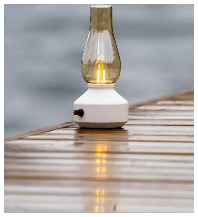 Lampada in stile lanterna accesa su un molo di legno bagnato, con il suo bagliore che si riflette sulle assi contro uno sfondo grigio sfocato.