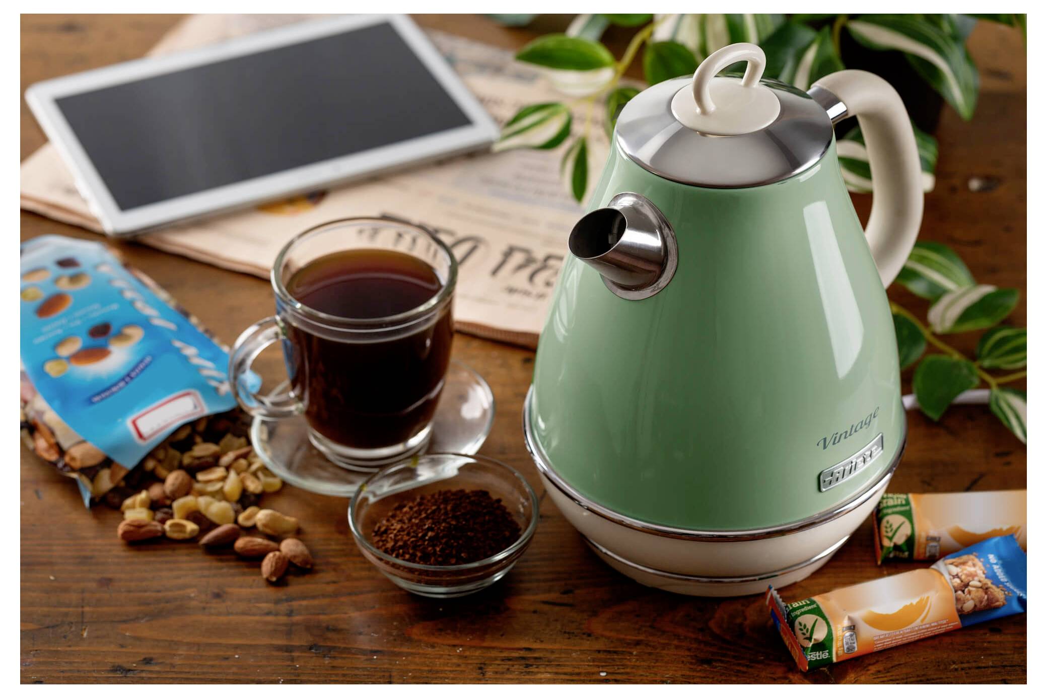 Bollitore elettrico verde accanto a un bicchiere di tè su un tavolo di legno con chicchi di caffè, tablet, noci e barrette di snack.