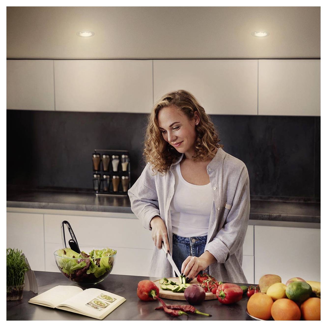Donna che prepara le verdure in una cucina moderna, affetta un cetriolo al bancone con ingredienti per insalata, un libro di ricette aperto e frutta fresca nelle vicinanze.