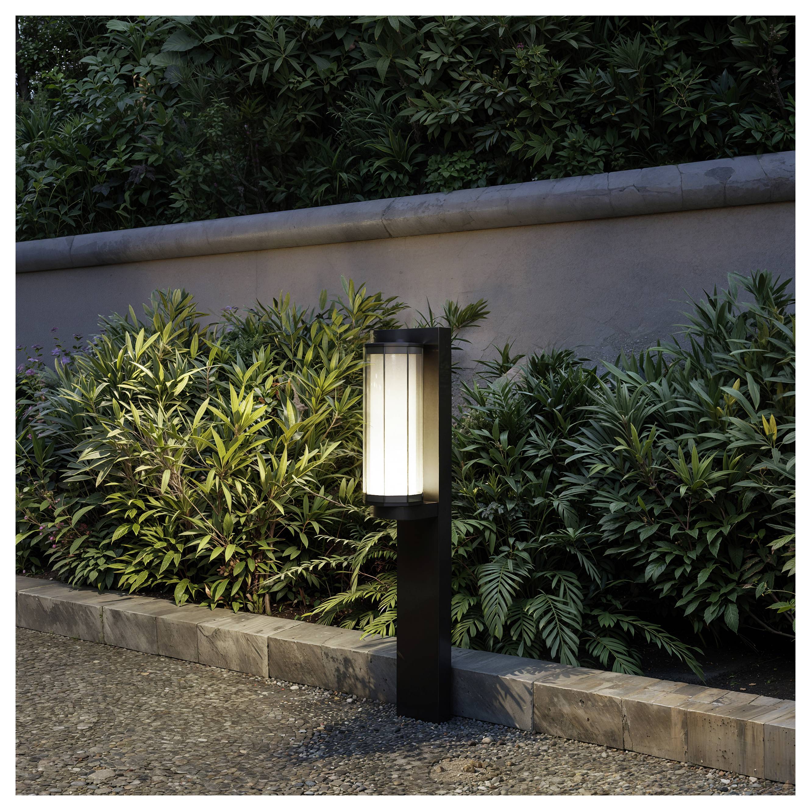 Luce da esterno nera a palo che brilla accanto a un'aiuola e un muro di cemento al crepuscolo.
