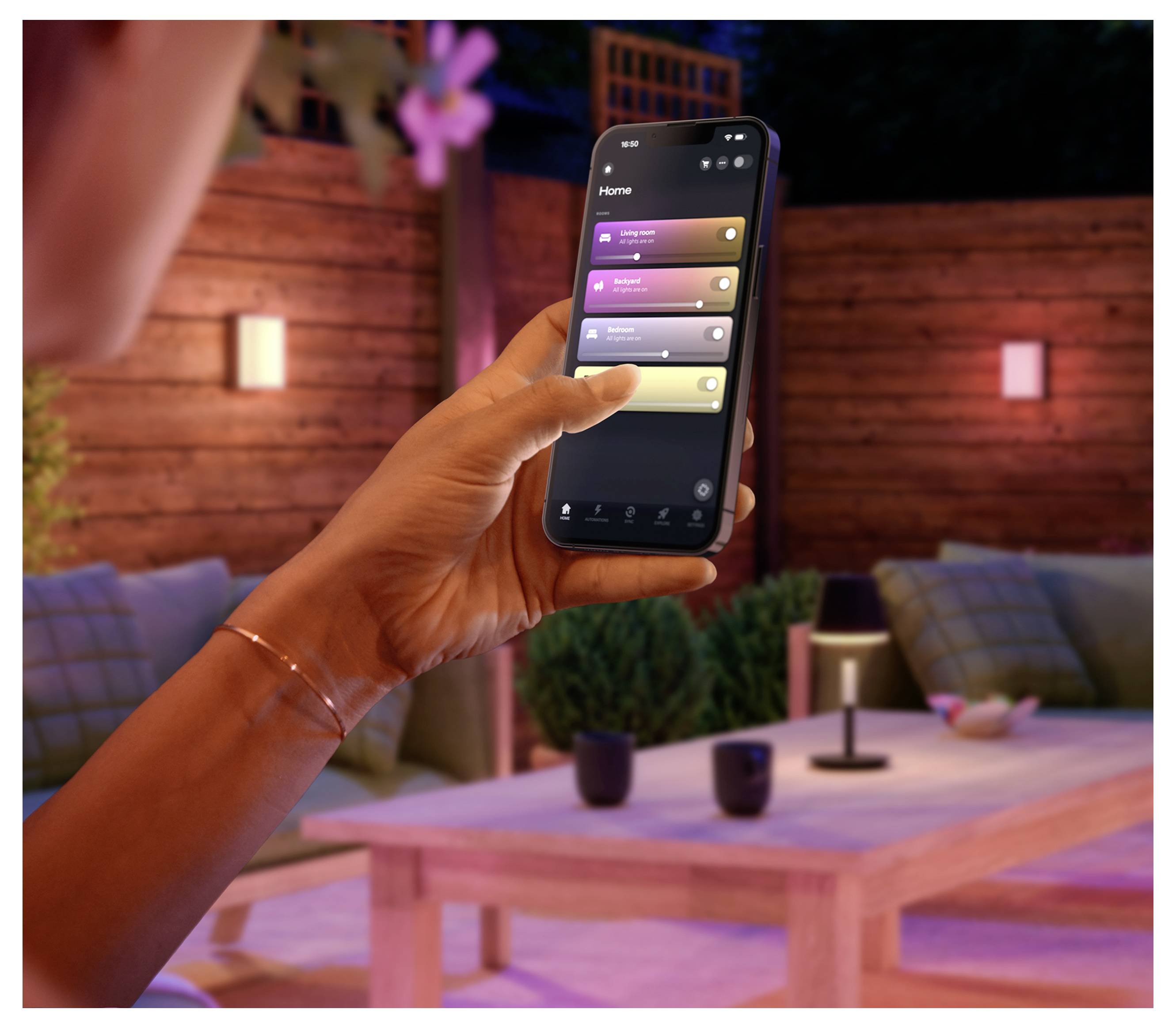 Una mano che tiene uno smartphone mostrando un'app per la casa con quattro cursori di controllo delle luci in un'accogliente e calda ambientazione di patio o salotto.
