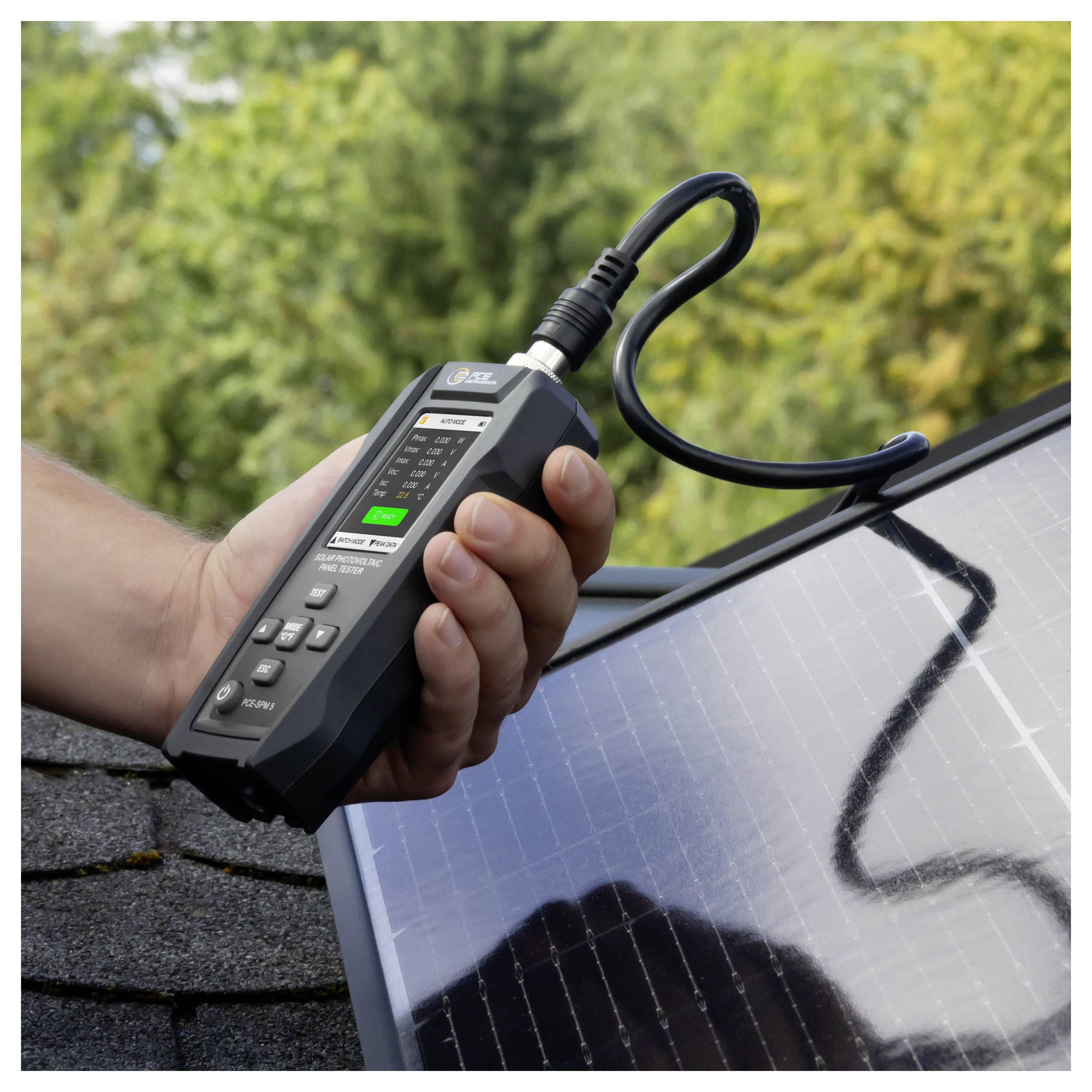 Una mano che tiene un tester per pannelli solari collegato tramite cavo a un pannello fotovoltaico all'aperto.