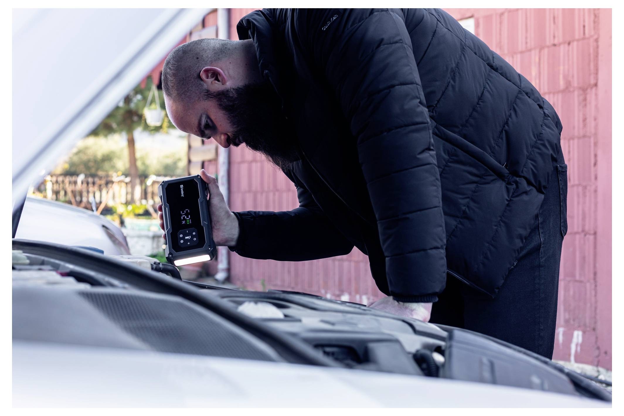 Un uomo ispeziona il motore di un'auto con un telefono portatile o uno strumento diagnostico mentre si sporge sul cofano aperto all'aperto.