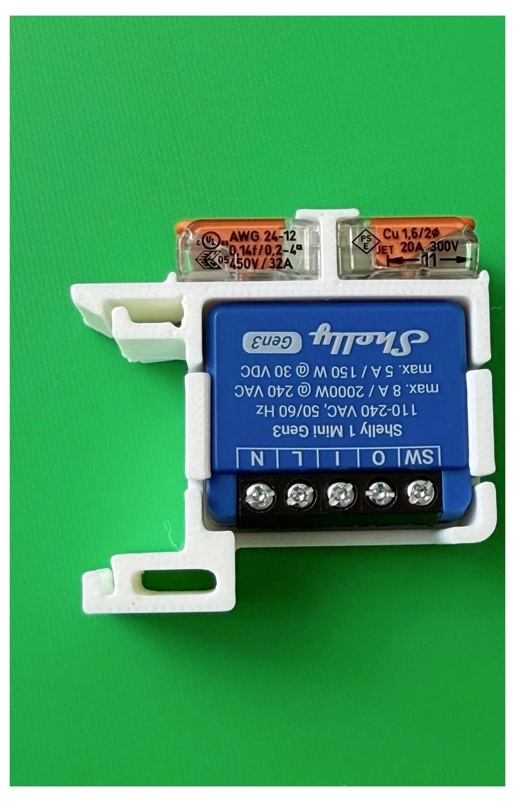 Primo piano di un modulo dimmer Shelly blu montato in una staffa di plastica bianca, con coperture terminali trasparenti, su uno sfondo verde.