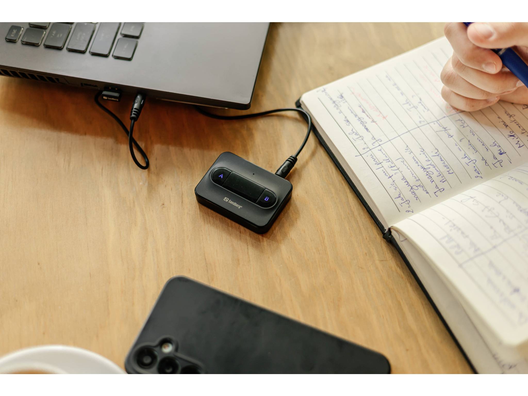 Una persona prende appunti accanto a un laptop e a degli auricolari wireless in carica su una scrivania di legno, con un telefono in primo piano.