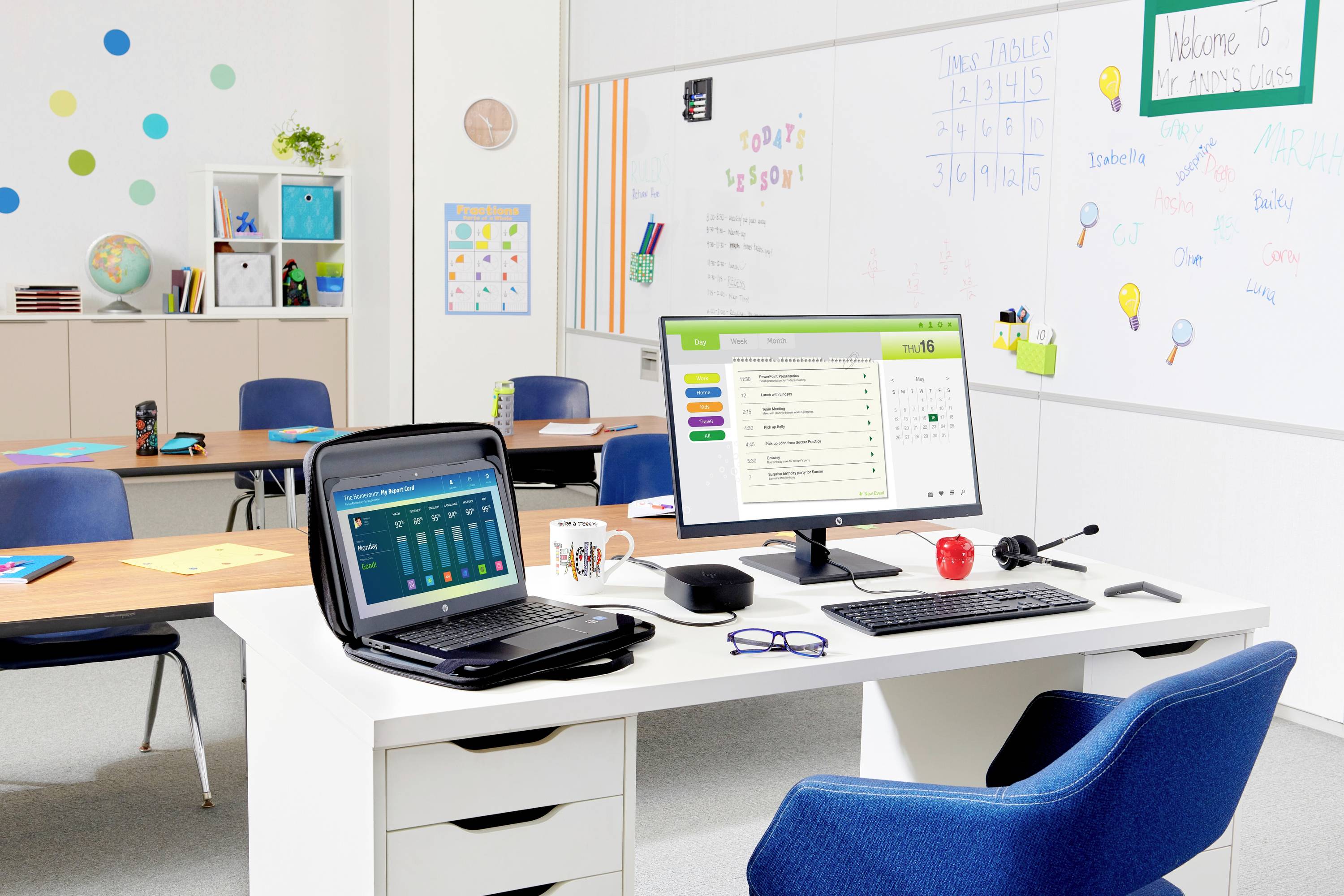 Aula vuota con una scrivania dell'insegnante, laptop e monitor, tastiera, tazza, occhiali e lavagne coperte di appunti e grafici.