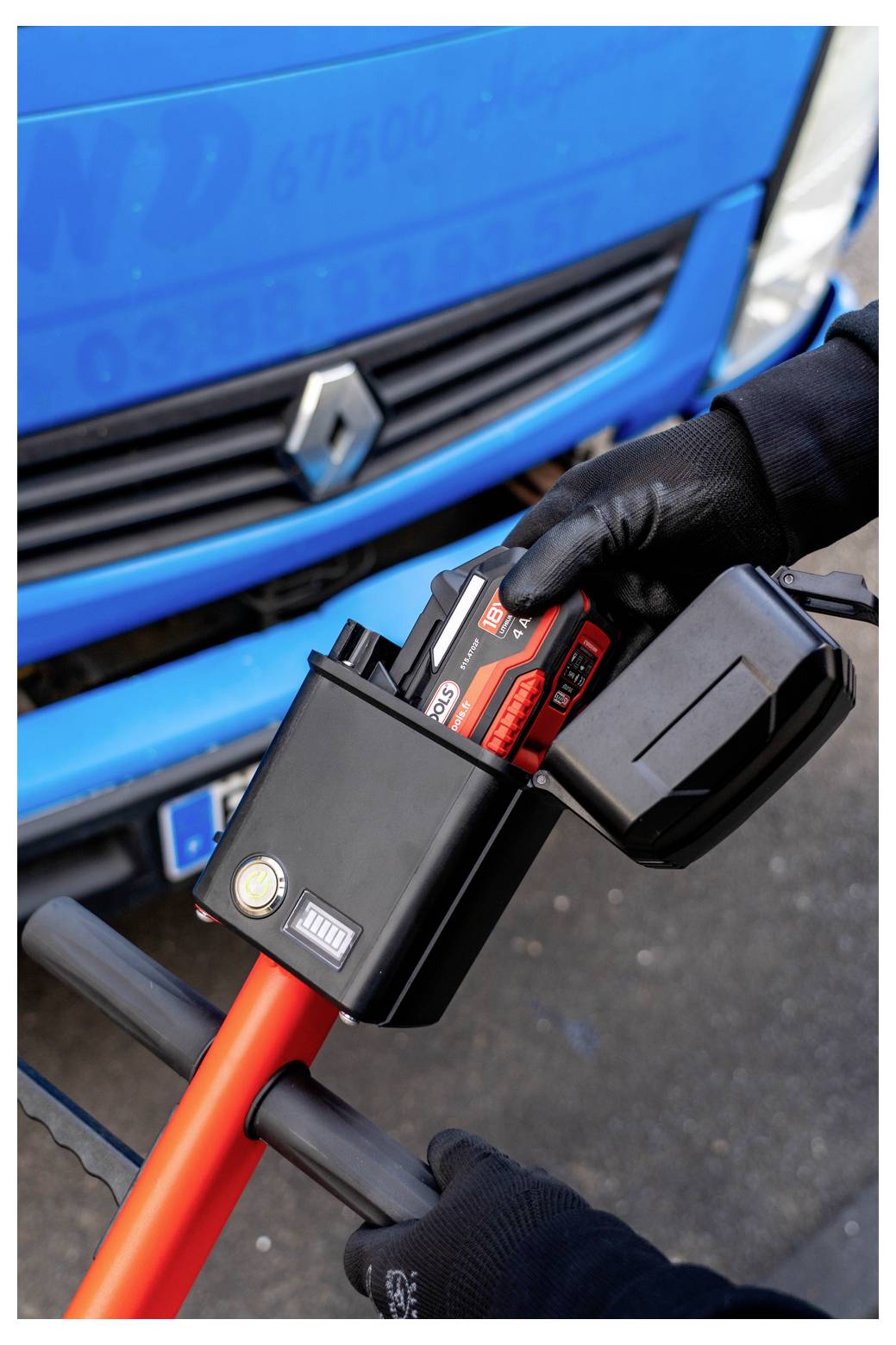 Le mani guantate inseriscono una batteria rossa in un caricabatterie montato su un veicolo di servizio Renault blu.