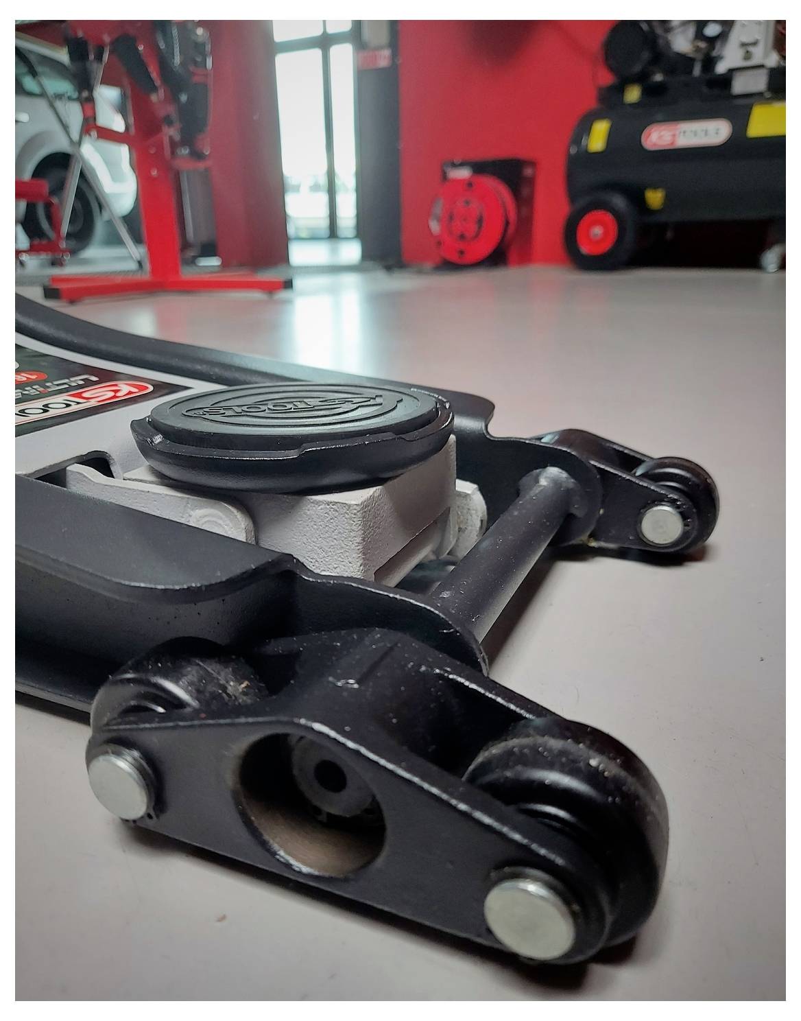 Primo piano di un accessorio per macchina da palestra su pavimento, che mostra un connettore di cavo metallico nero e un braccio pivot in una sala fitness rossa.