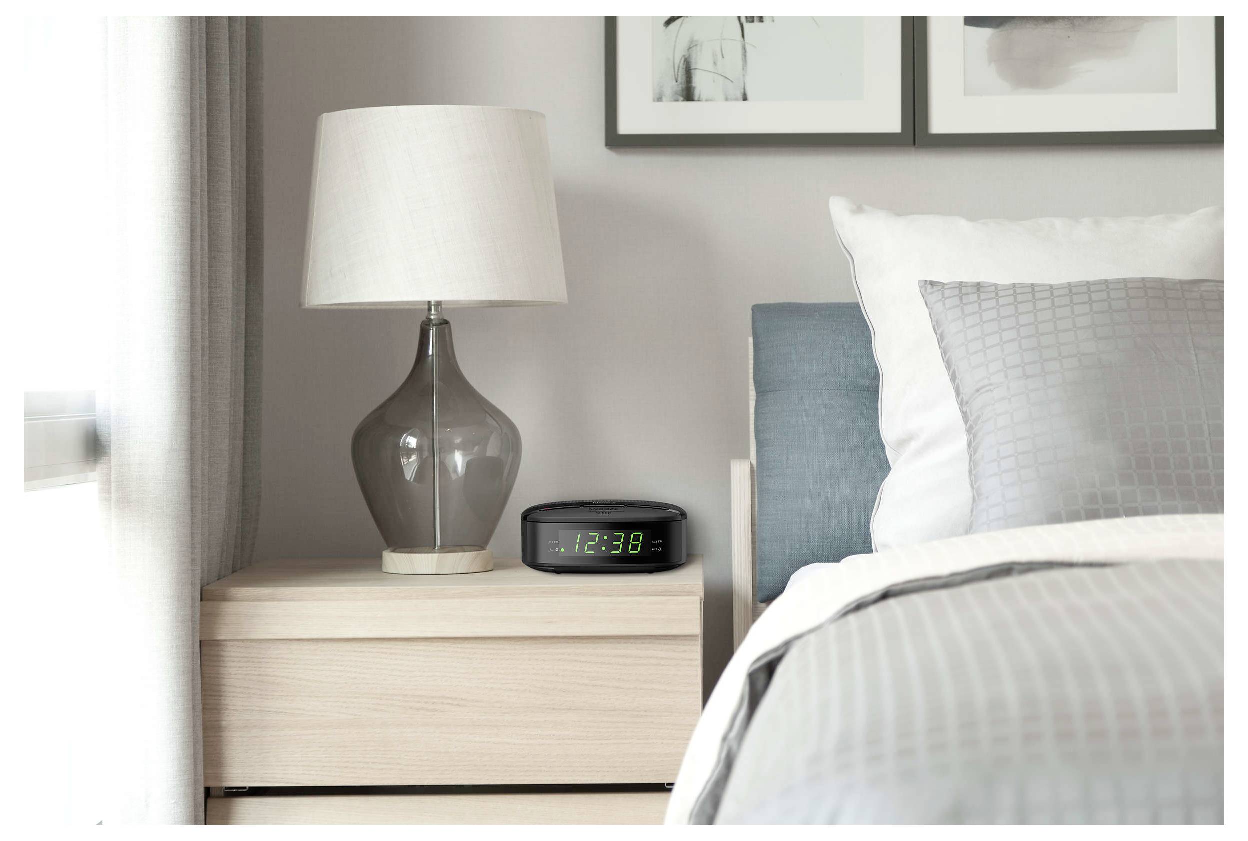 Comodino della camera da letto con lampada e sveglia digitale che mostra le 12:38 accanto a un letto con biancheria bianca e grigia.