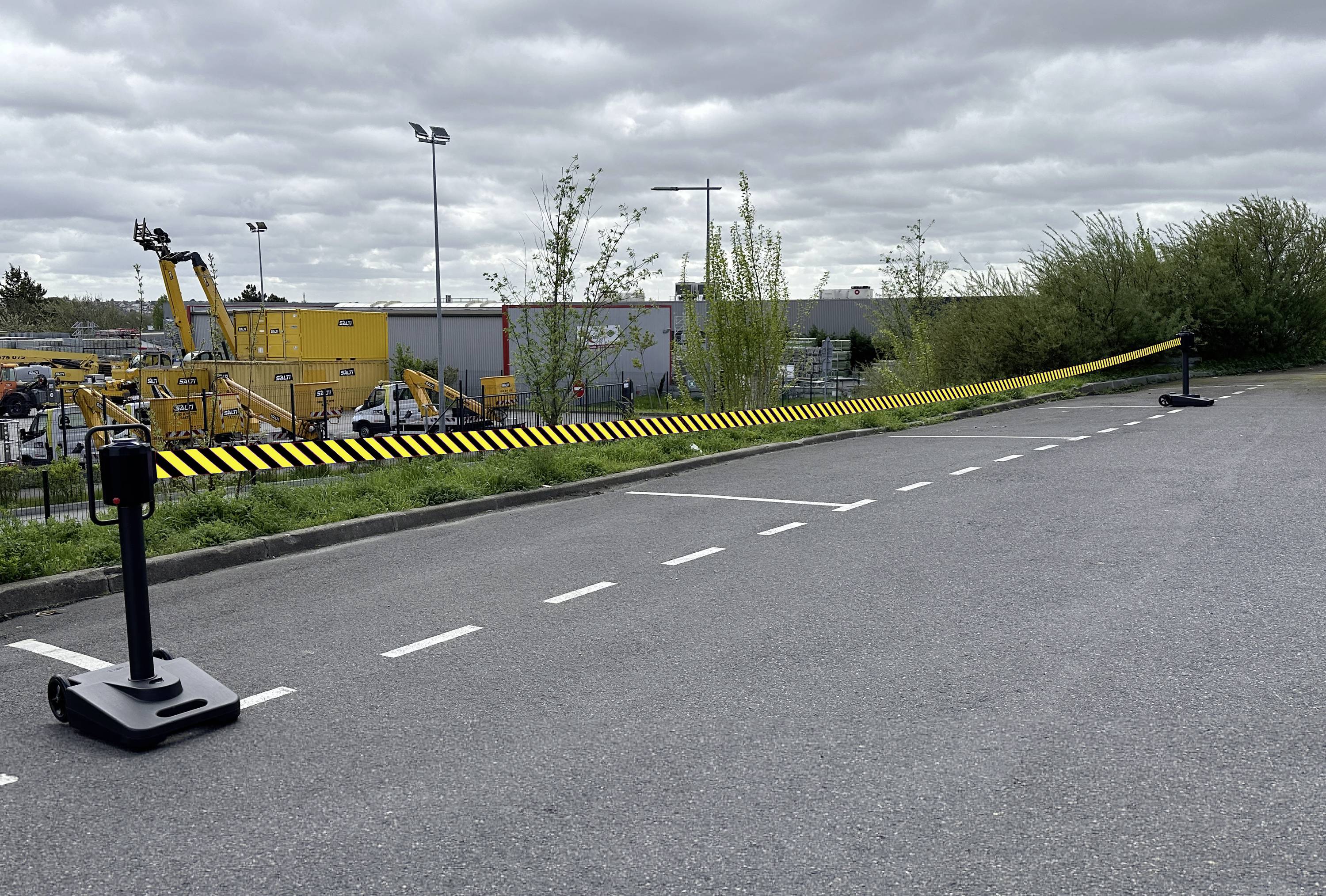 Nastro di avvertimento giallo e nero blocca un'area di lavoro lungo la strada con attrezzature da costruzione e edifici industriali sotto cieli nuvolosi.