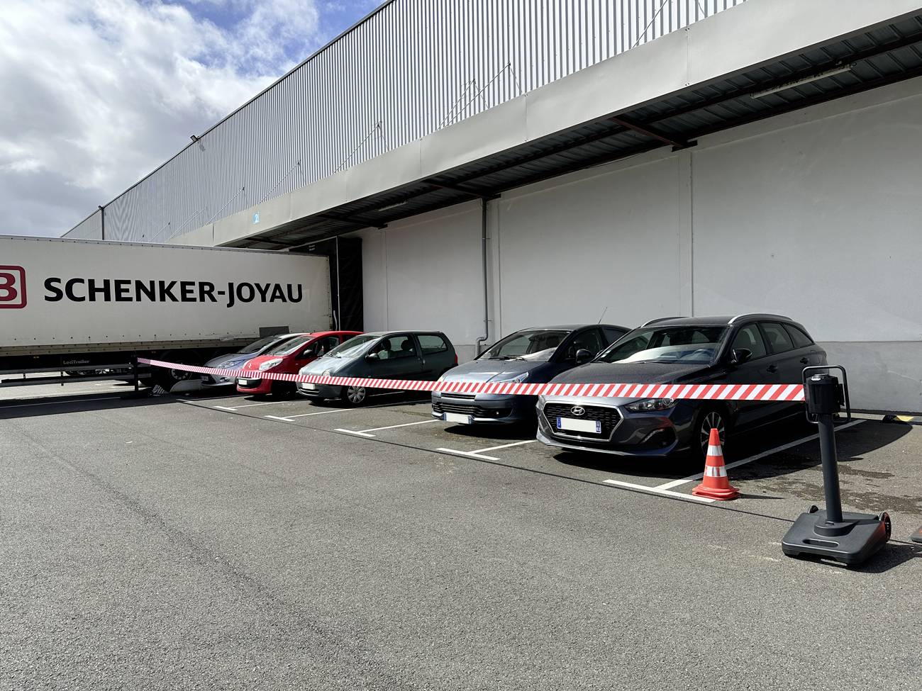Quattro auto parcheggiate accanto a un rimorchio Schenker-Joyau in un'area industriale, bloccate da nastro di barriera rosso e bianco e da un cono di traffico.