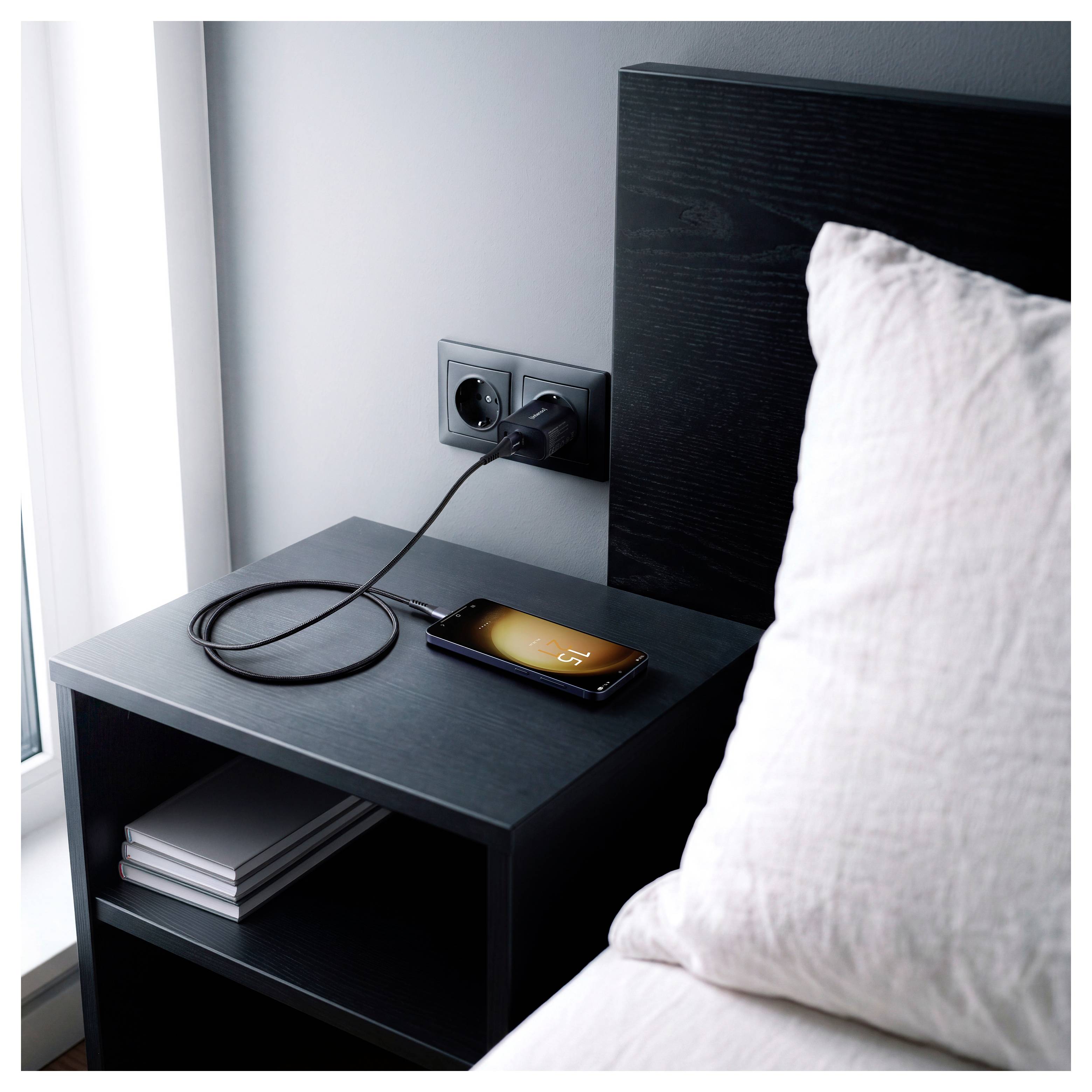 Uno smartphone in carica su un comodino accanto a un letto, con il cavo collegato a una presa a muro e il telefono che visualizza il 15%
