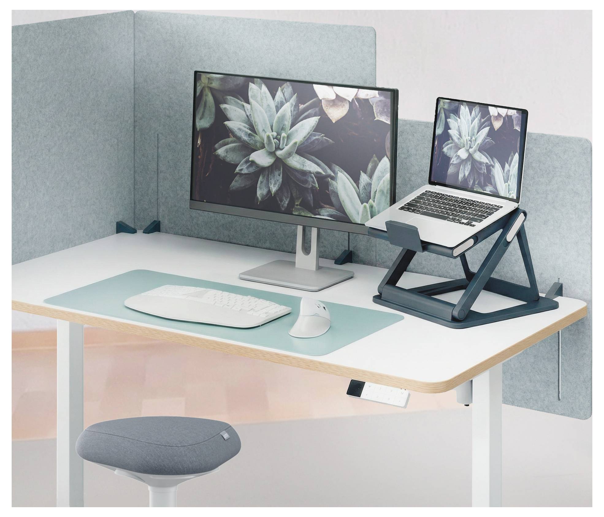 Configurazione minimalista della scrivania con monitor, laptop su supporto, tastiera, mouse e sgabello in un angolo ufficio dai toni pastello.