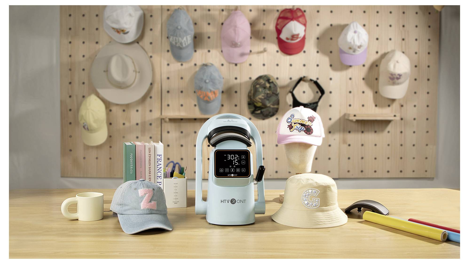 Una pressa termica HTVRONT portatile blu chiaro si trova su un tavolo da lavoro, circondata da cappelli decorati e forniture artigianali in uno spazio di lavoro per la personalizzazione di cappelli.