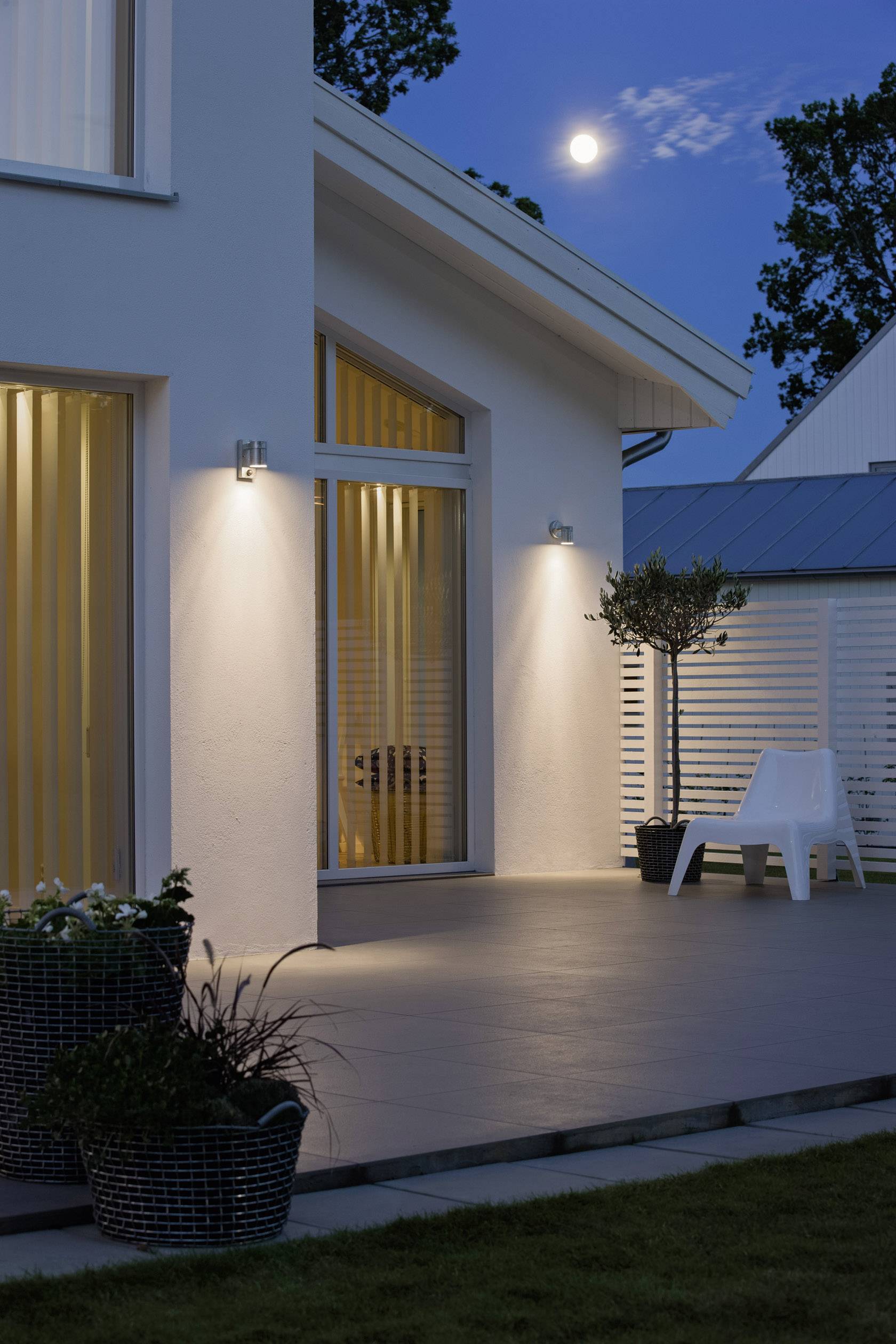 Una casa moderna con terrazza di notte, illuminata da luci esterne. La luna piena splende nel cielo. Su un'unica sedia posta sulla terrazza.