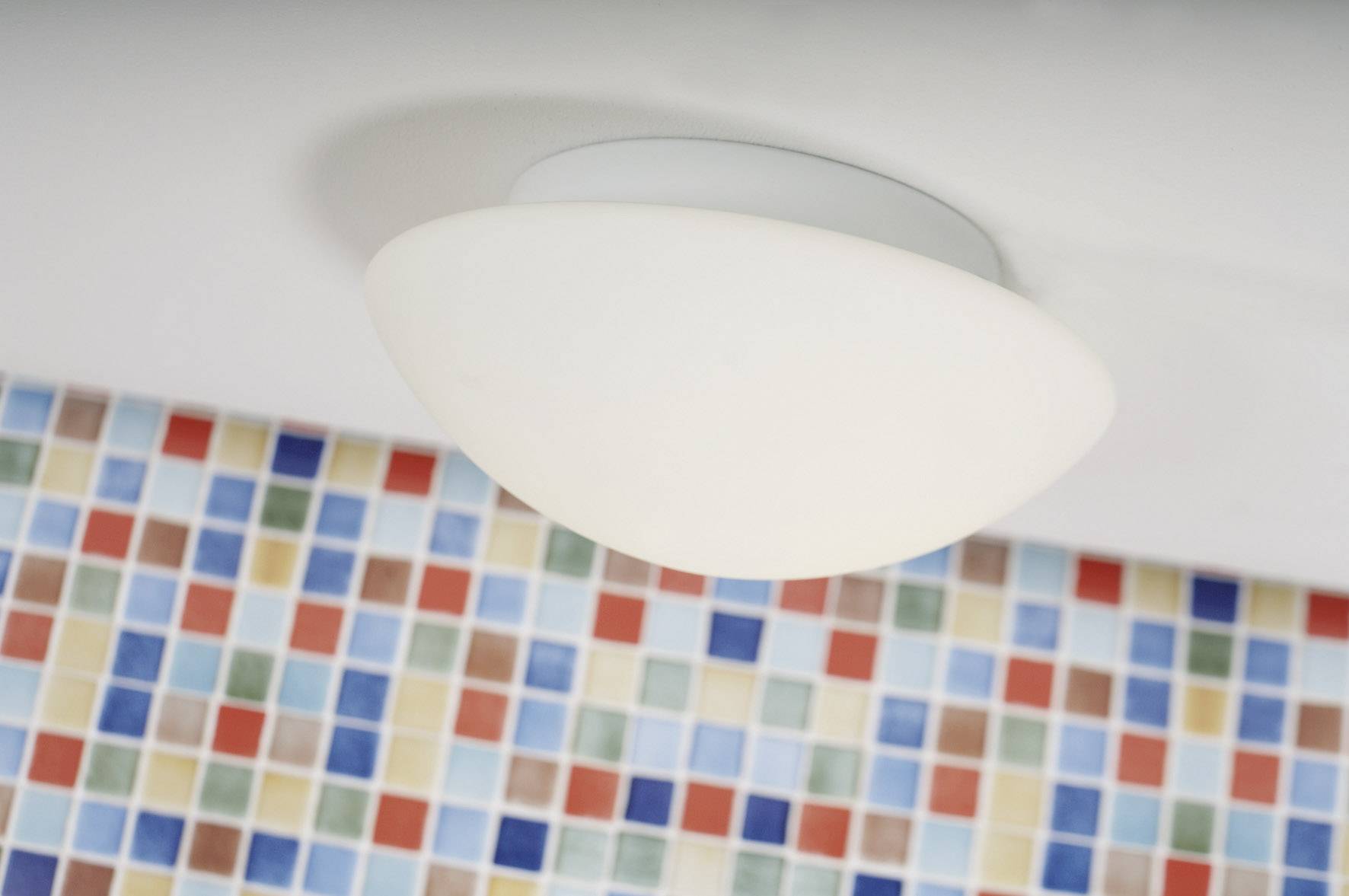Plafoniera a forma di semisfera sul soffitto, sotto di essa un muro rivestito di piastrelle mosaicate quadrate e colorate.