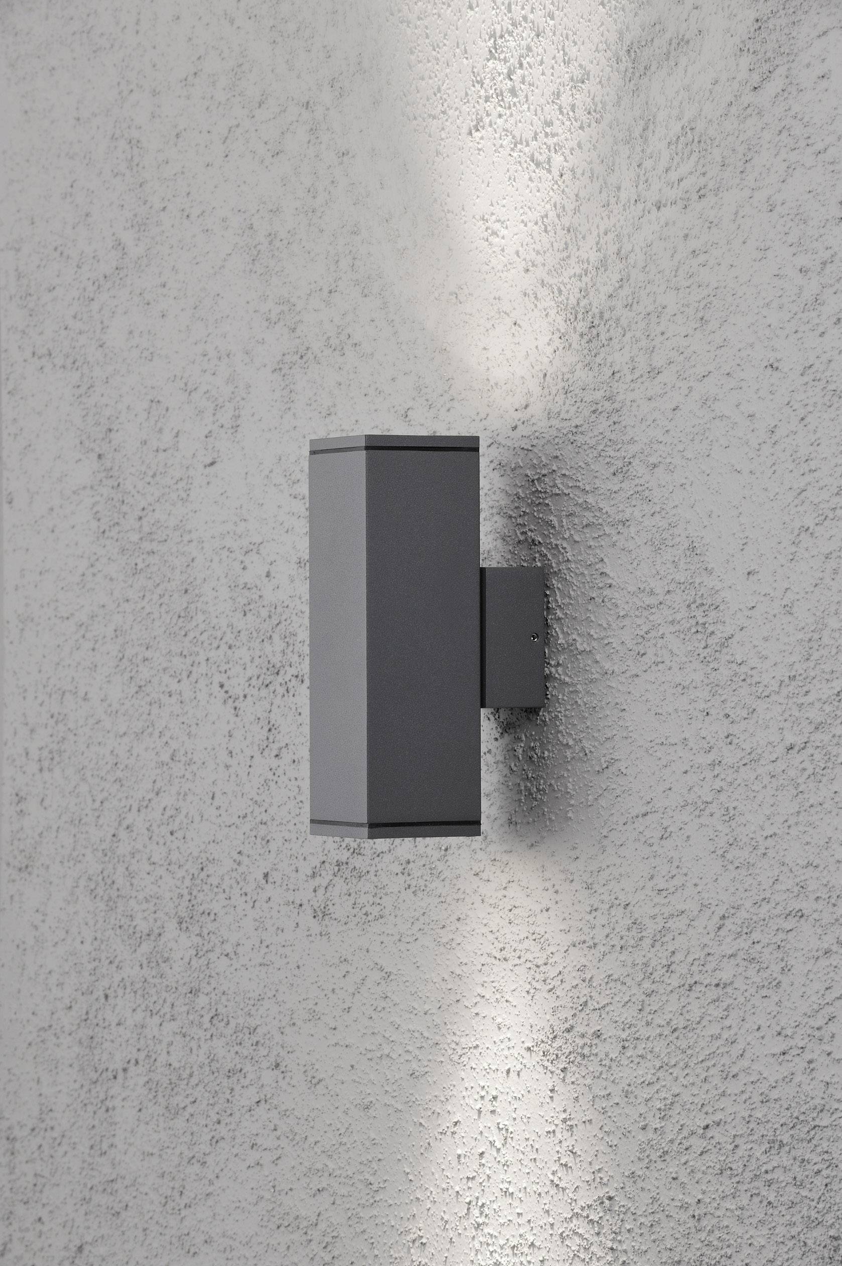 Un applique moderno grigio emana luce verso l'alto e verso il basso su una parete bianca strutturata.