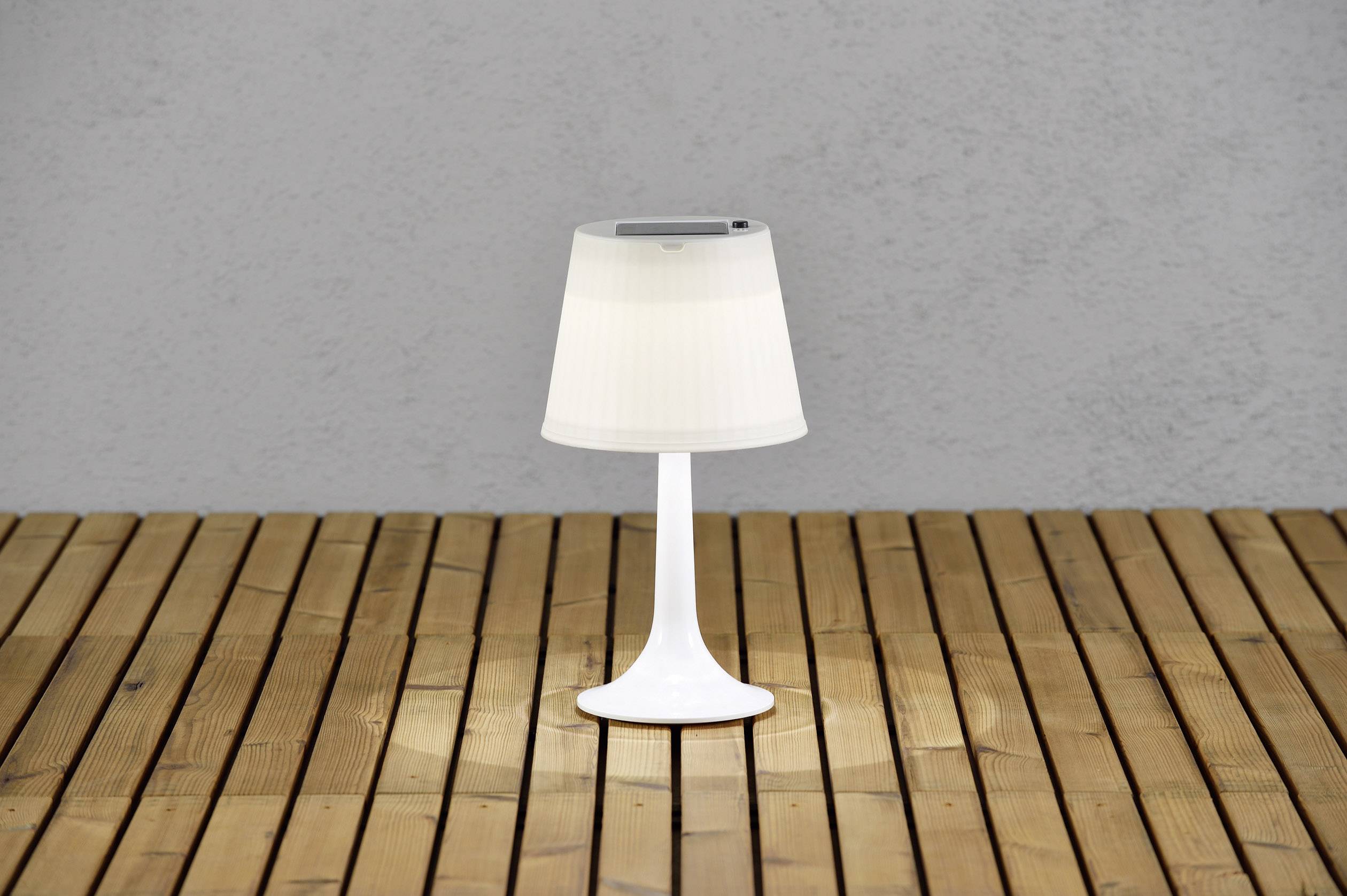 Lampada da tavolo bianca su un pavimento esterno in legno, illumina una piccola area sottostante. Sullo sfondo un muro di cemento grigio.