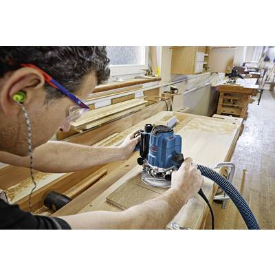 Un uomo lavora in un laboratorio con una fresatrice blu su una tavola di legno, indossa occhiali di protezione e tappi per le orecchie.