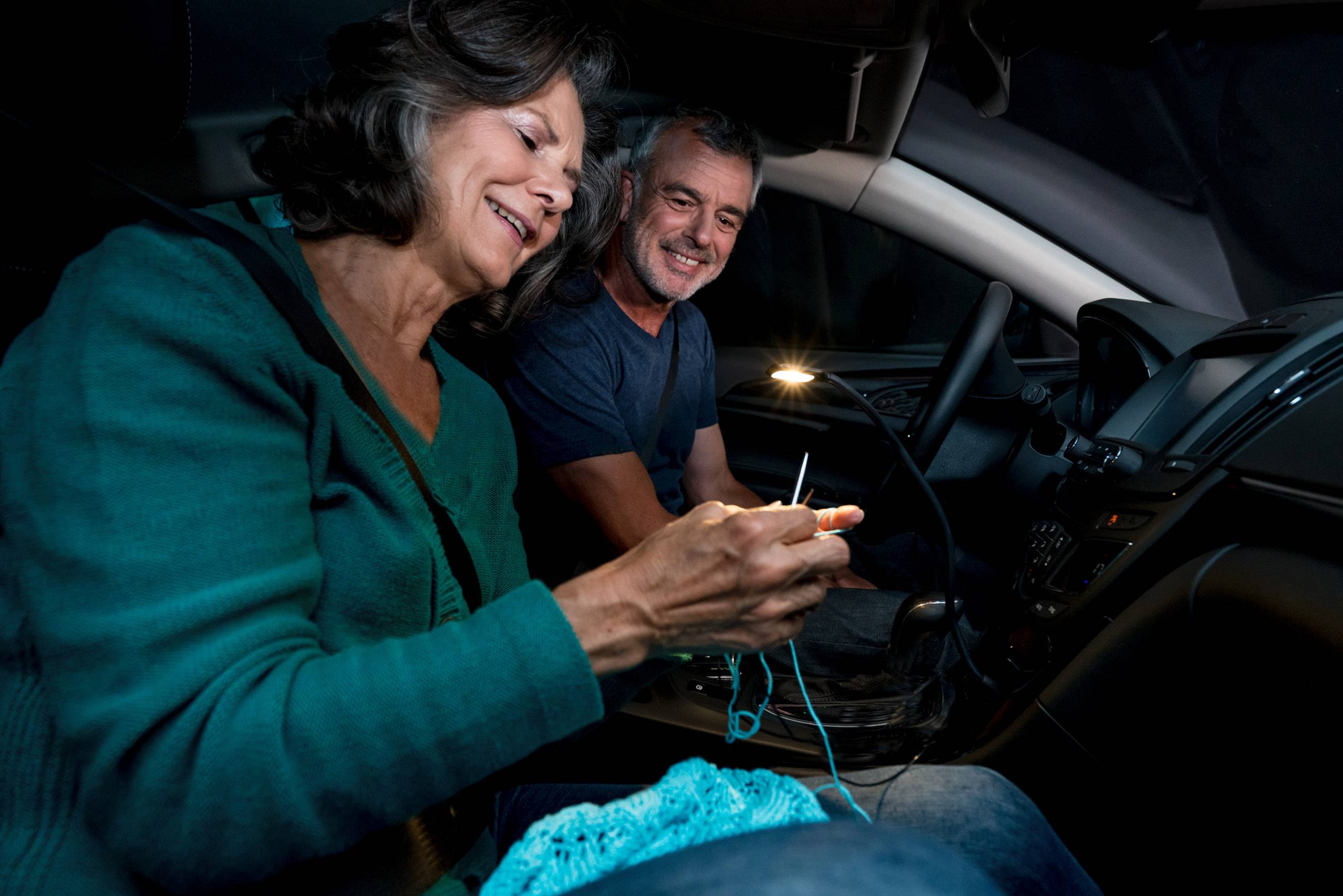 Una donna anziana lavora a maglia in auto sotto la luce di lettura, mentre un uomo sorridente la osserva. L'atmosfera appare accogliente e rilassata.