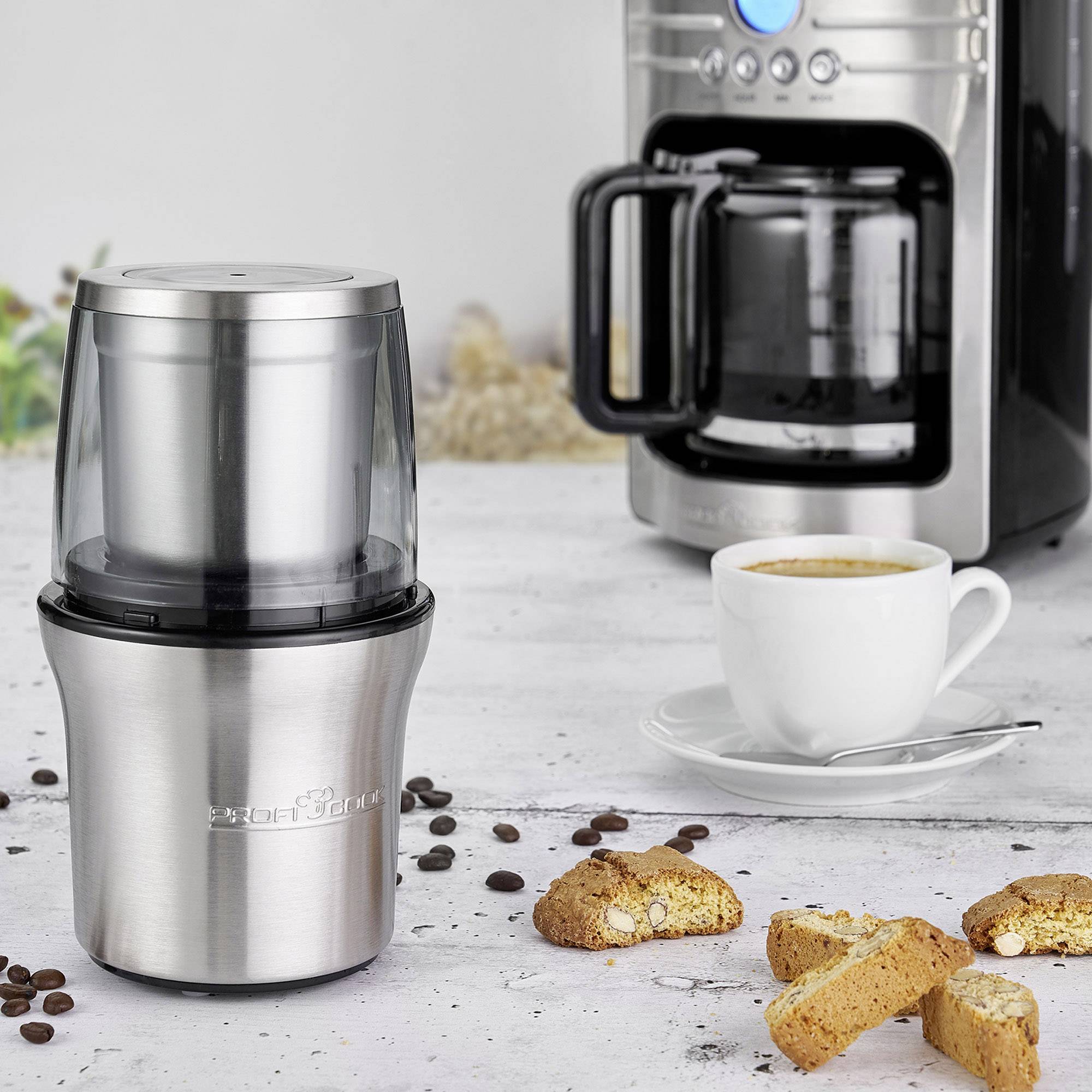 Un contenitore elettrico per macinare il caffè è posizionato su un tavolo accanto a una tazza di caffè, una macchina da caffè e alcuni biscotti.