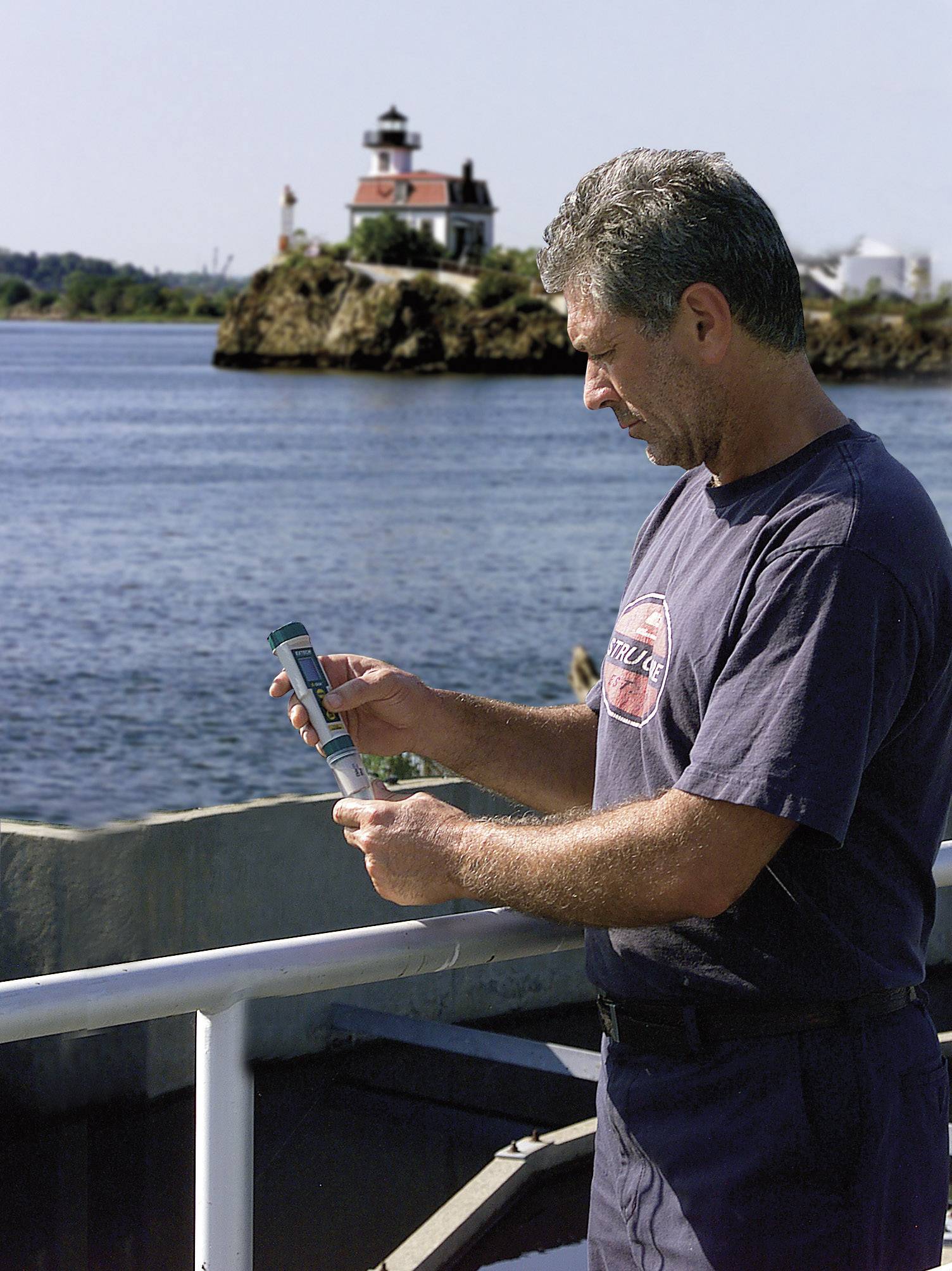 Un uomo è in piedi su una barca e osserva un oggetto tra le sue mani. Sullo sfondo è visibile un faro su una piccola isola.