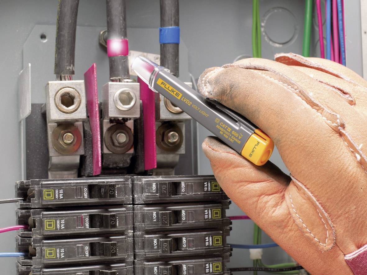 Mano con guanto che tiene un tester su un circuito elettrico. Fusibili e cavi sono visibili. Verifica di sicurezza.