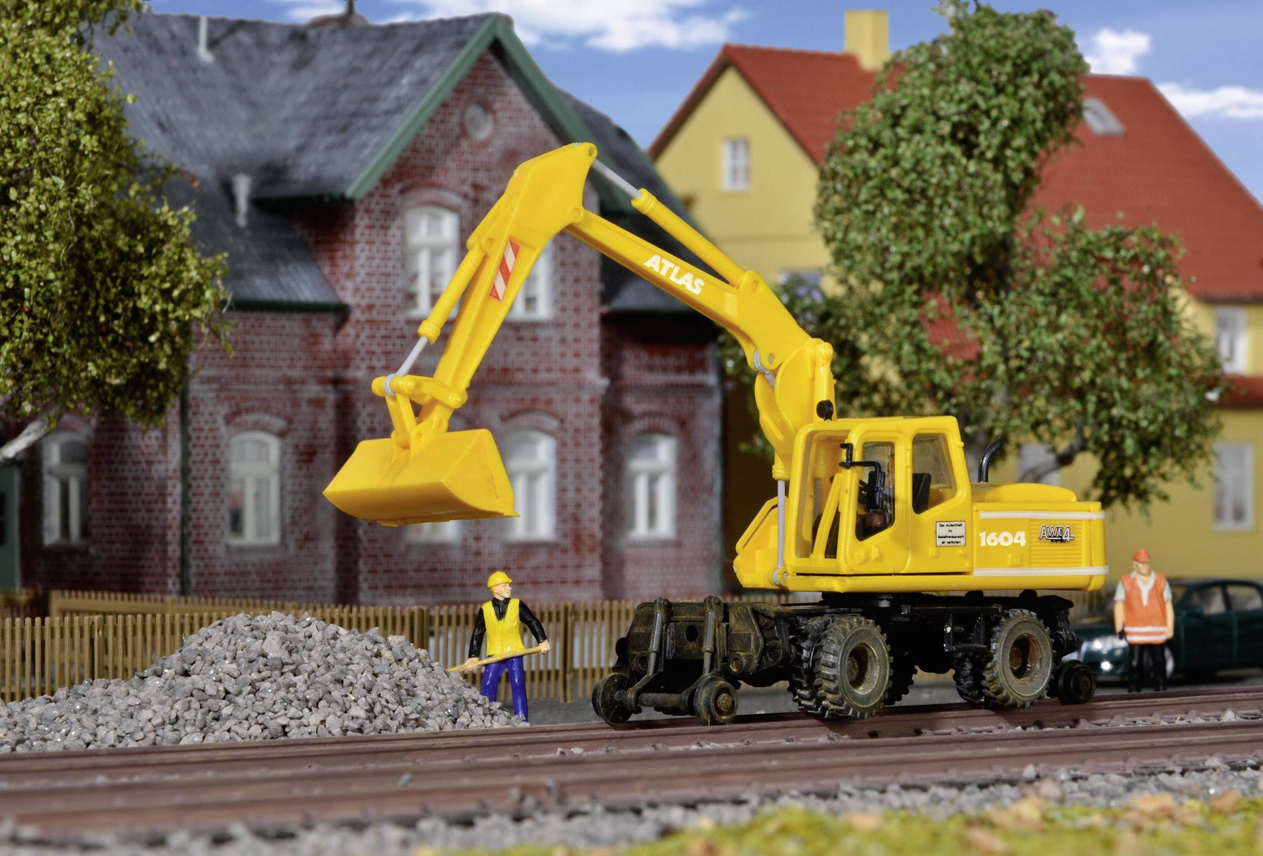 Un escavatore giallo solleva pietre in un cantiere edile. Due operai sono in piedi accanto, con un grande edificio rosso-marrone sullo sfondo.