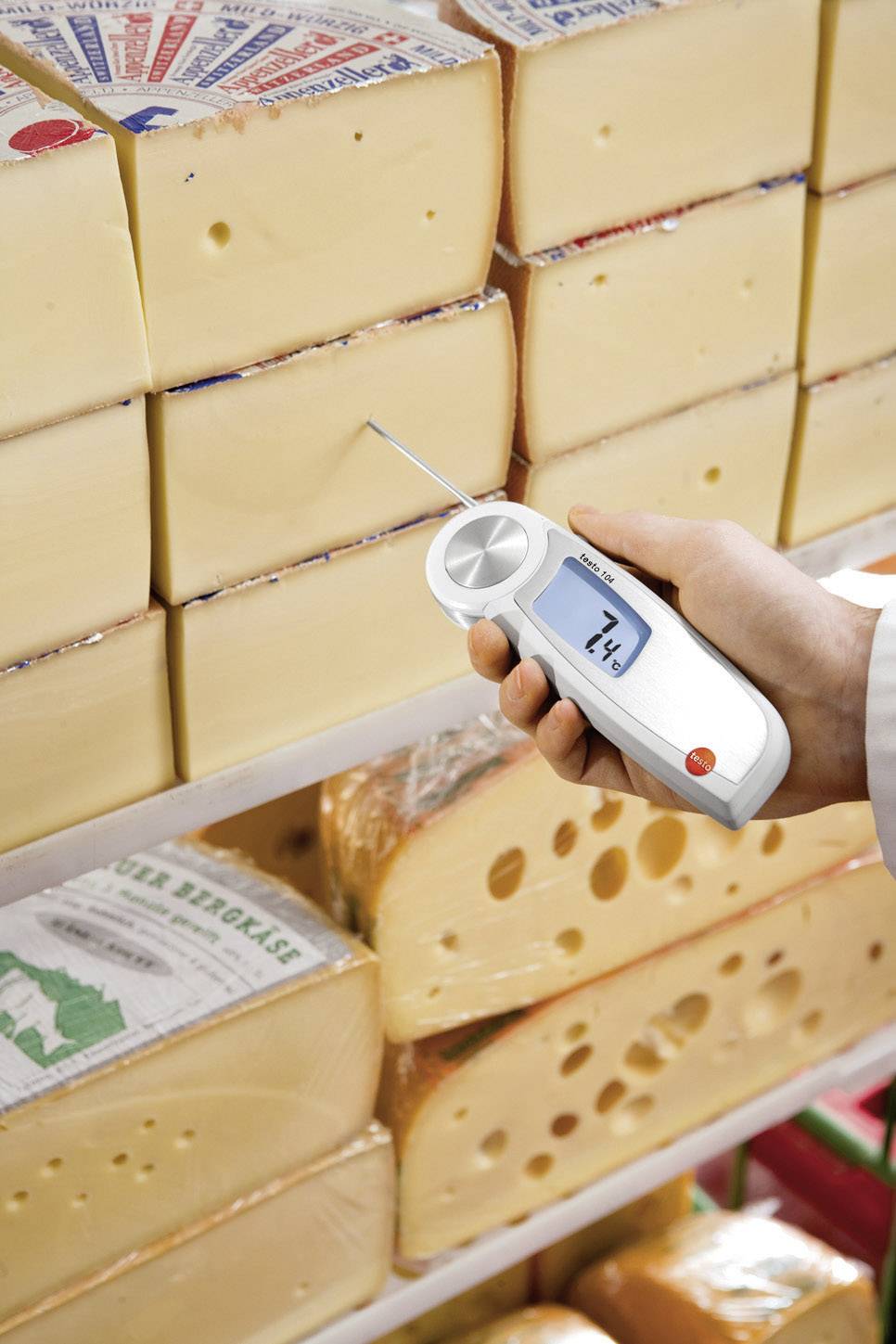 Una persona misura la temperatura di una forma di formaggio nel magazzino utilizzando un termometro digitale. Il display mostra 14,4 gradi.