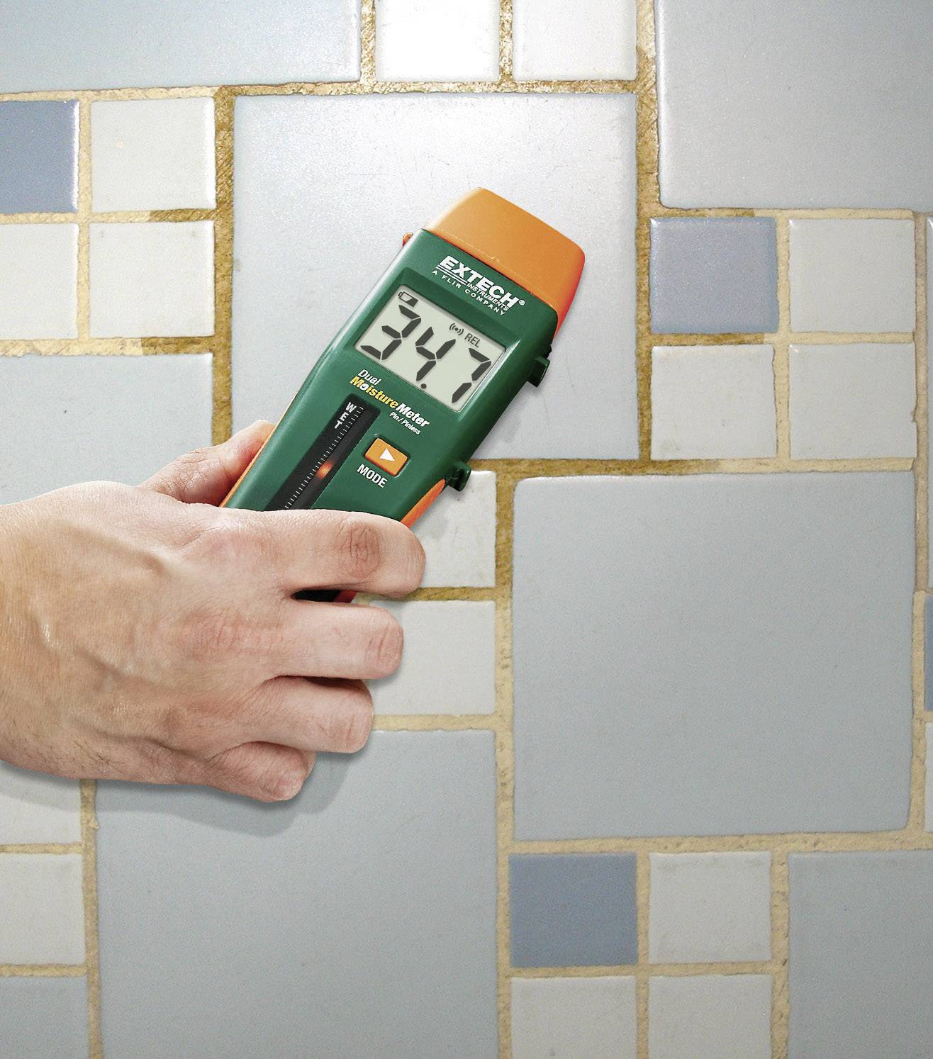 Una mano tiene un misuratore di umidità contro un muro piastrellato blu, mostrando il 34,7 per cento.