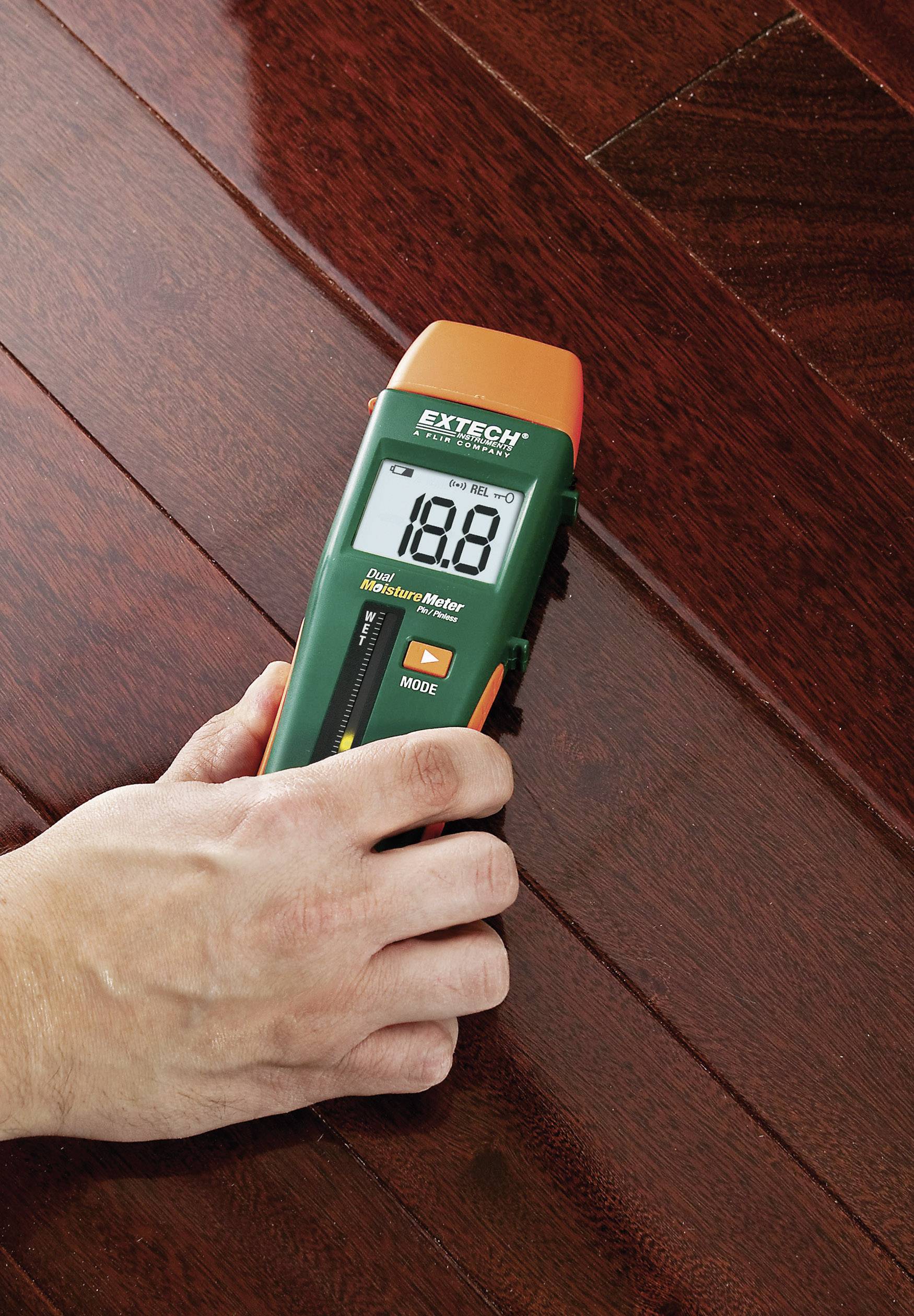 Una mano tiene un misuratore di umidità su un pavimento di legno scuro. Il display mostra '18,8'.