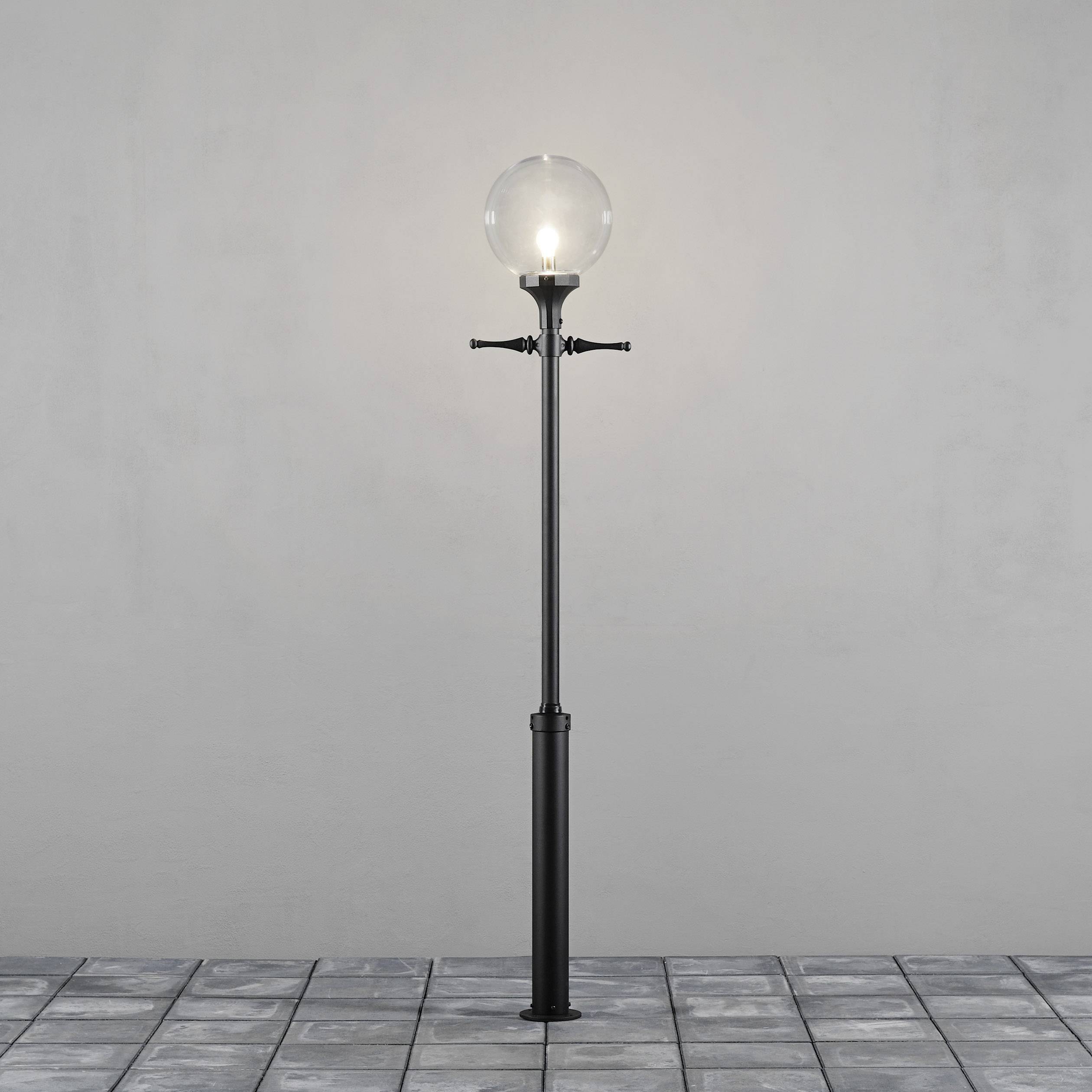 Una lampione nero con una lampadina sferica e trasparente su un pavimento lastricato davanti a un muro grigio.