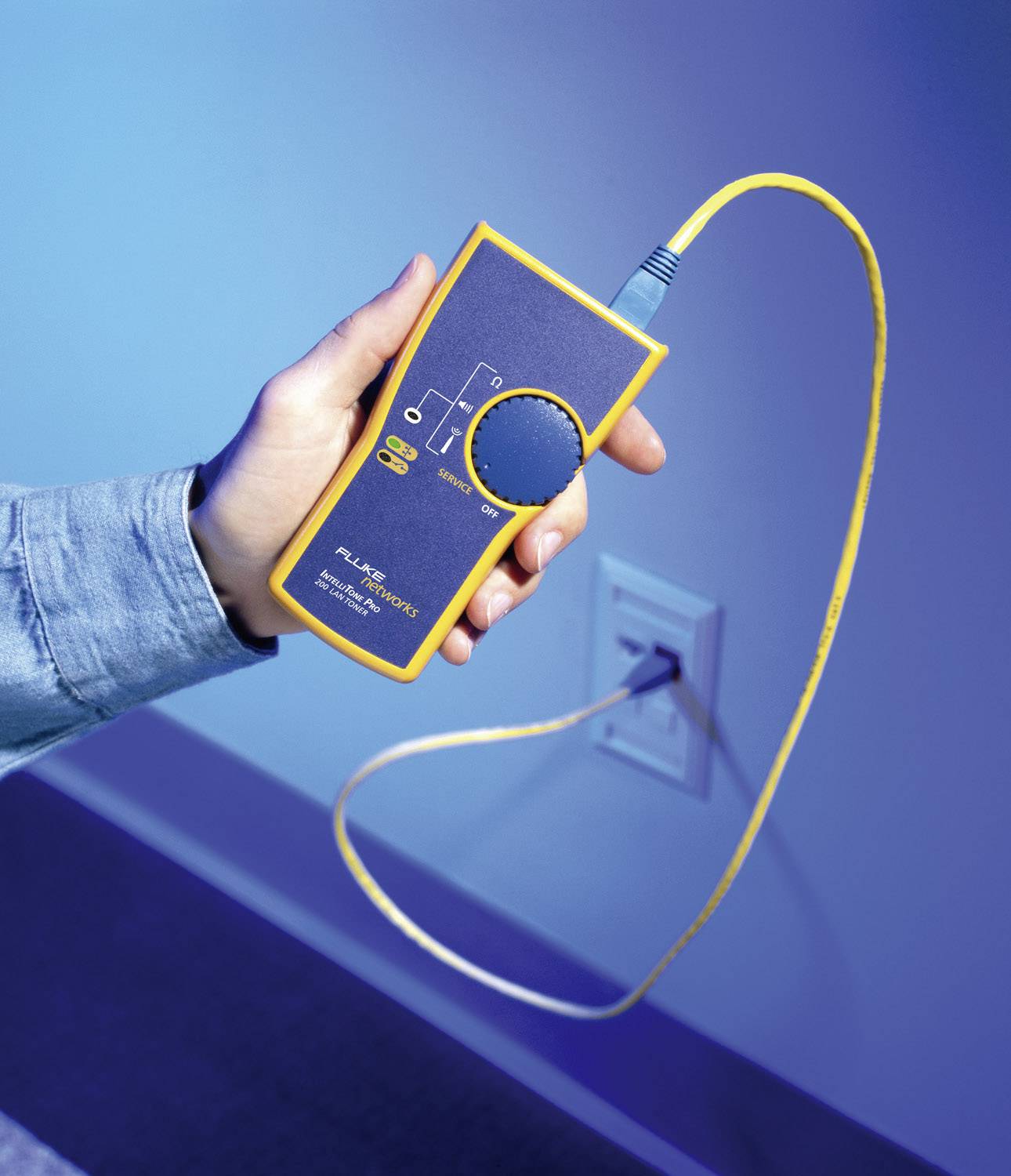 Una mano tiene un dispositivo elettrico giallo per misurazioni, con un cavo collegato a una presa a muro.