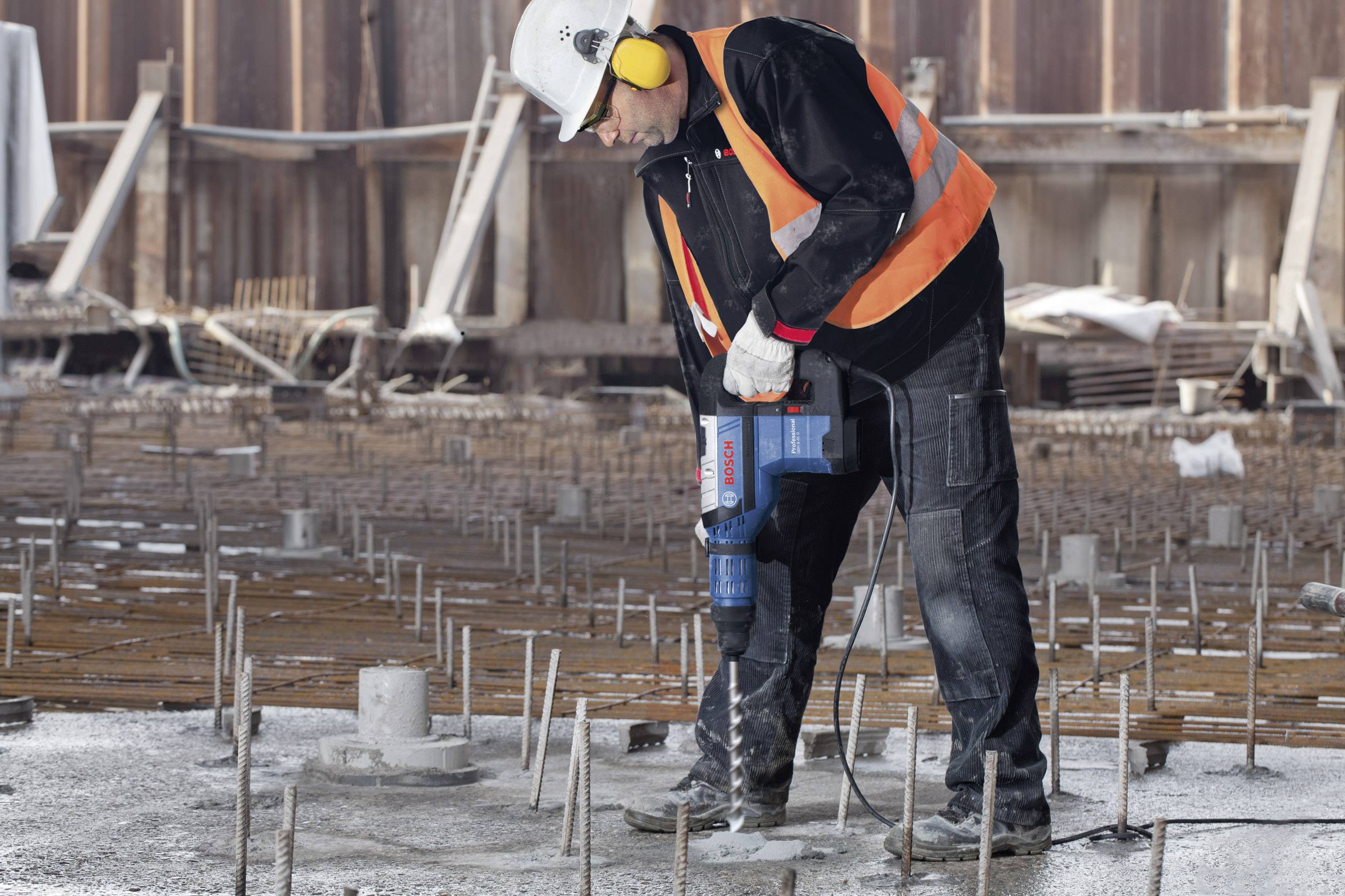 Un operaio edile con casco di protezione e protezioni per l'udito perfora una lastra di cemento con un trapano elettrico in un cantiere.