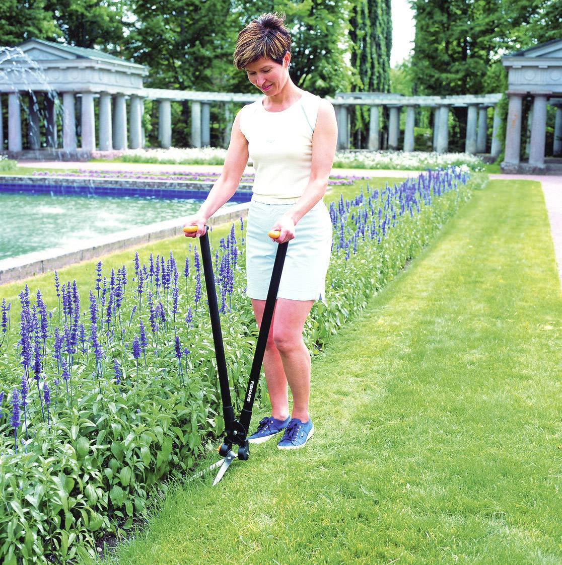 Una donna taglia il bordo di un'aiuola con un tagliasiepi lungo in un giardino curato con fontana e colonne.
