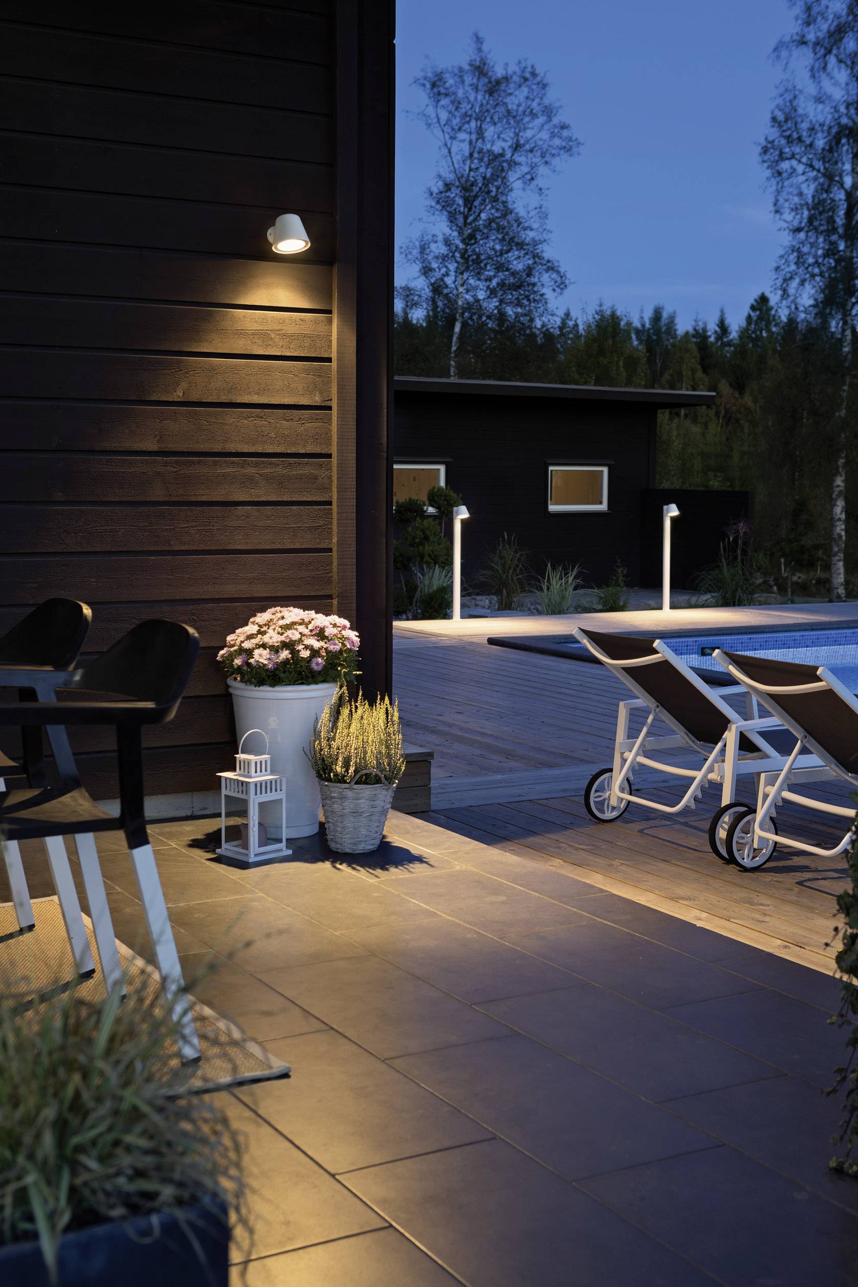 Terrazza al crepuscolo con illuminazione, sdraio e piante. La piscina è visibile sullo sfondo, circondata da alberi.