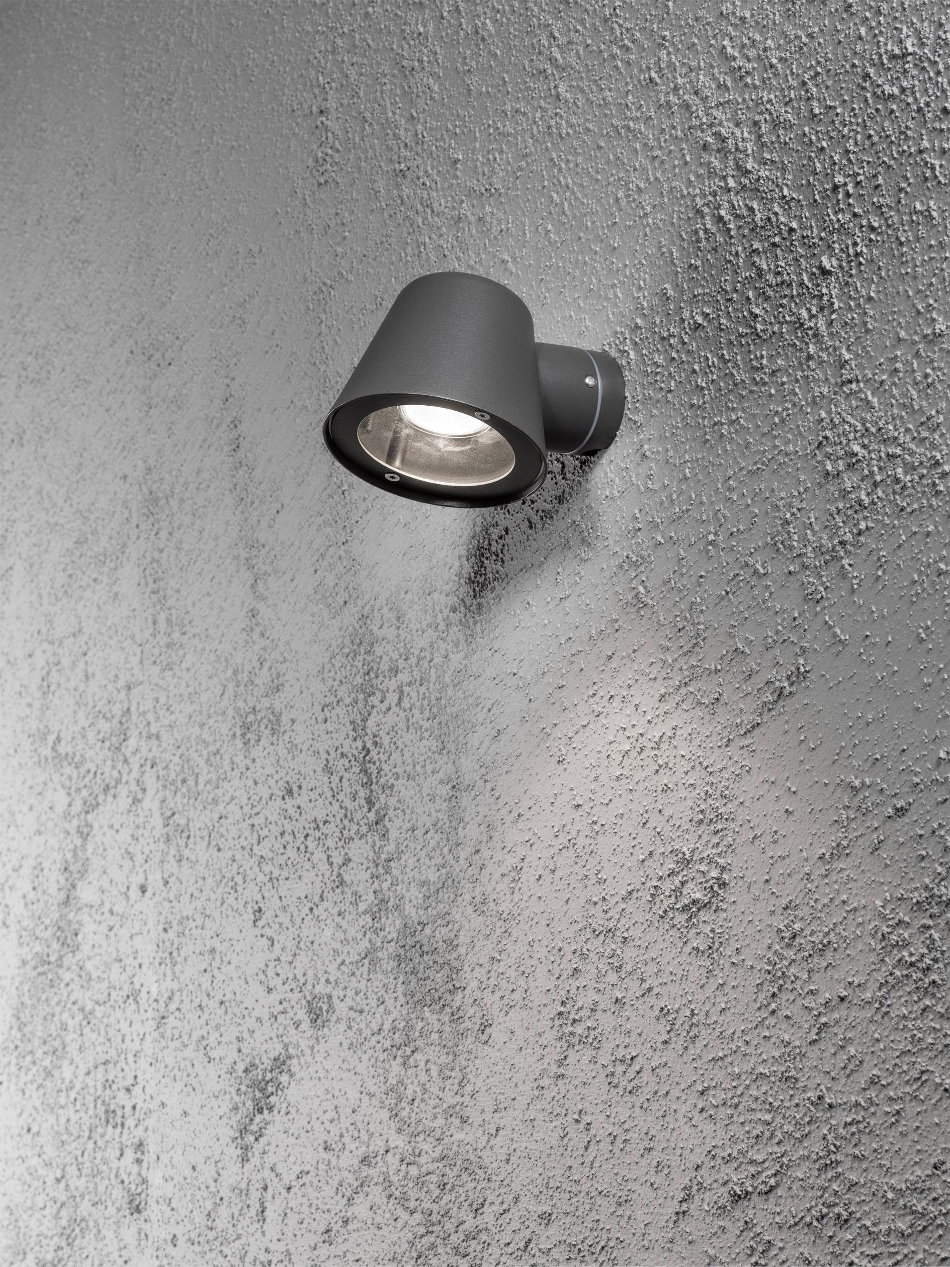 Una moderna lampada esterna nera su un muro di cemento strutturato, che illumina verso il basso offrendo una distribuzione uniforme della luce.