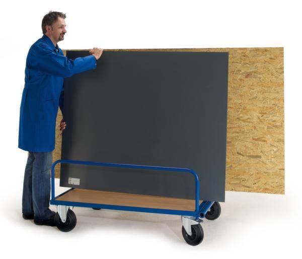 Un uomo con un camice blu sta caricando grandi lastre di legno e metallo su un carrello di trasporto blu.
