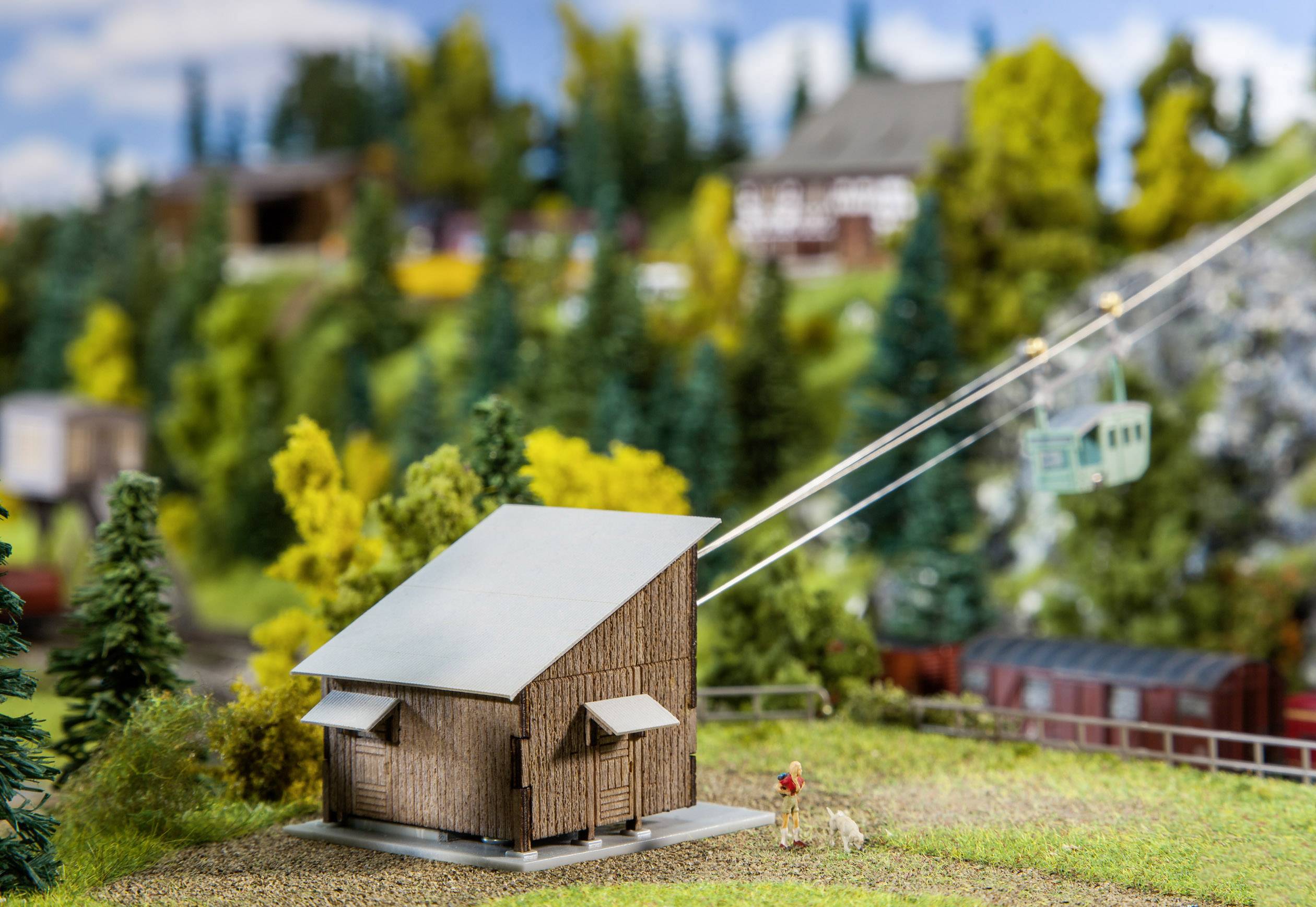 Modellino in miniatura di un paesaggio rurale con una casetta di legno, una funivia e alberi sullo sfondo che rappresentano una scena montana.