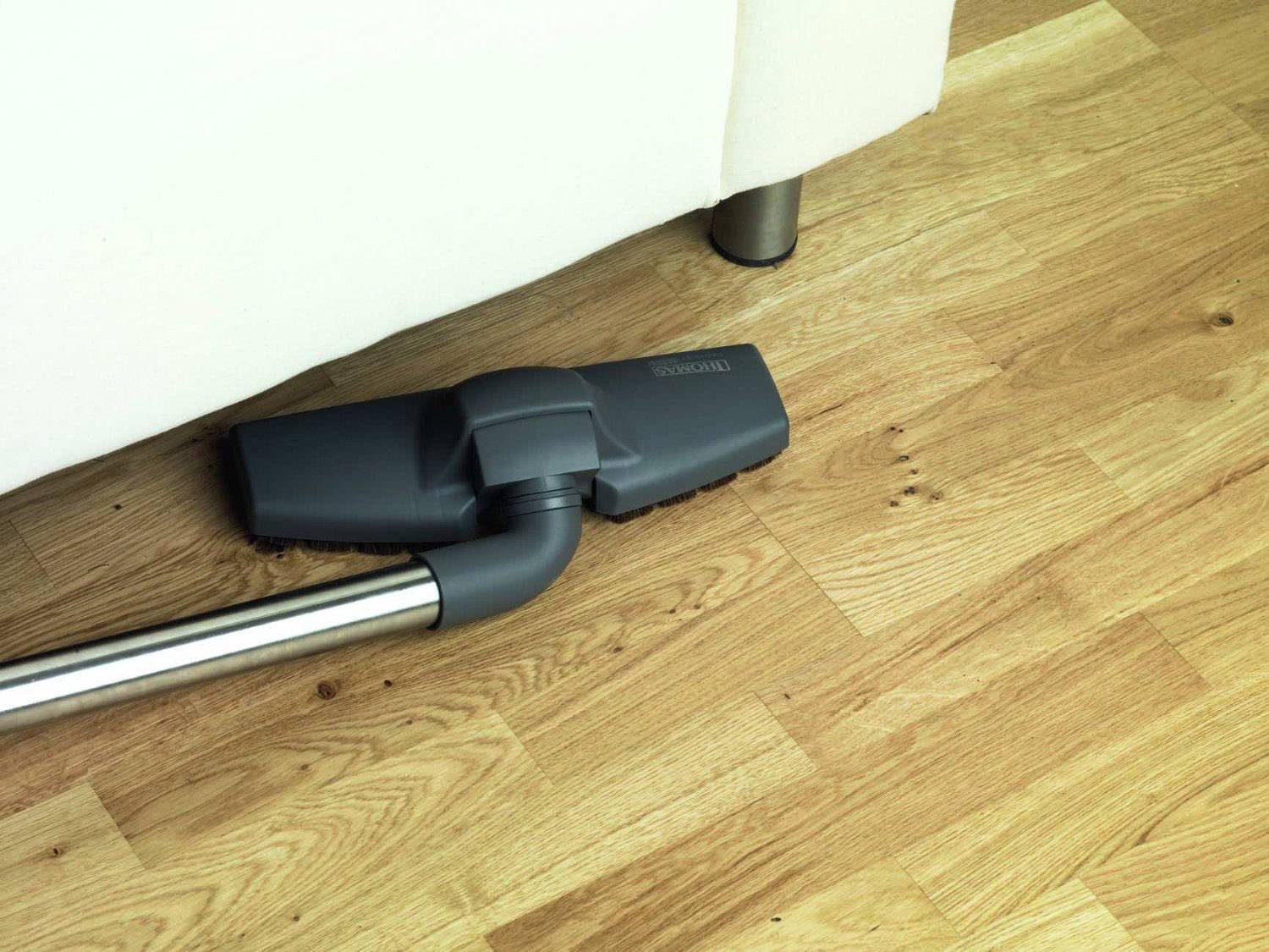 Un aspirapolvere pulisce il pavimento in legno sotto un divano bianco per rimuovere polvere e sporcizia.