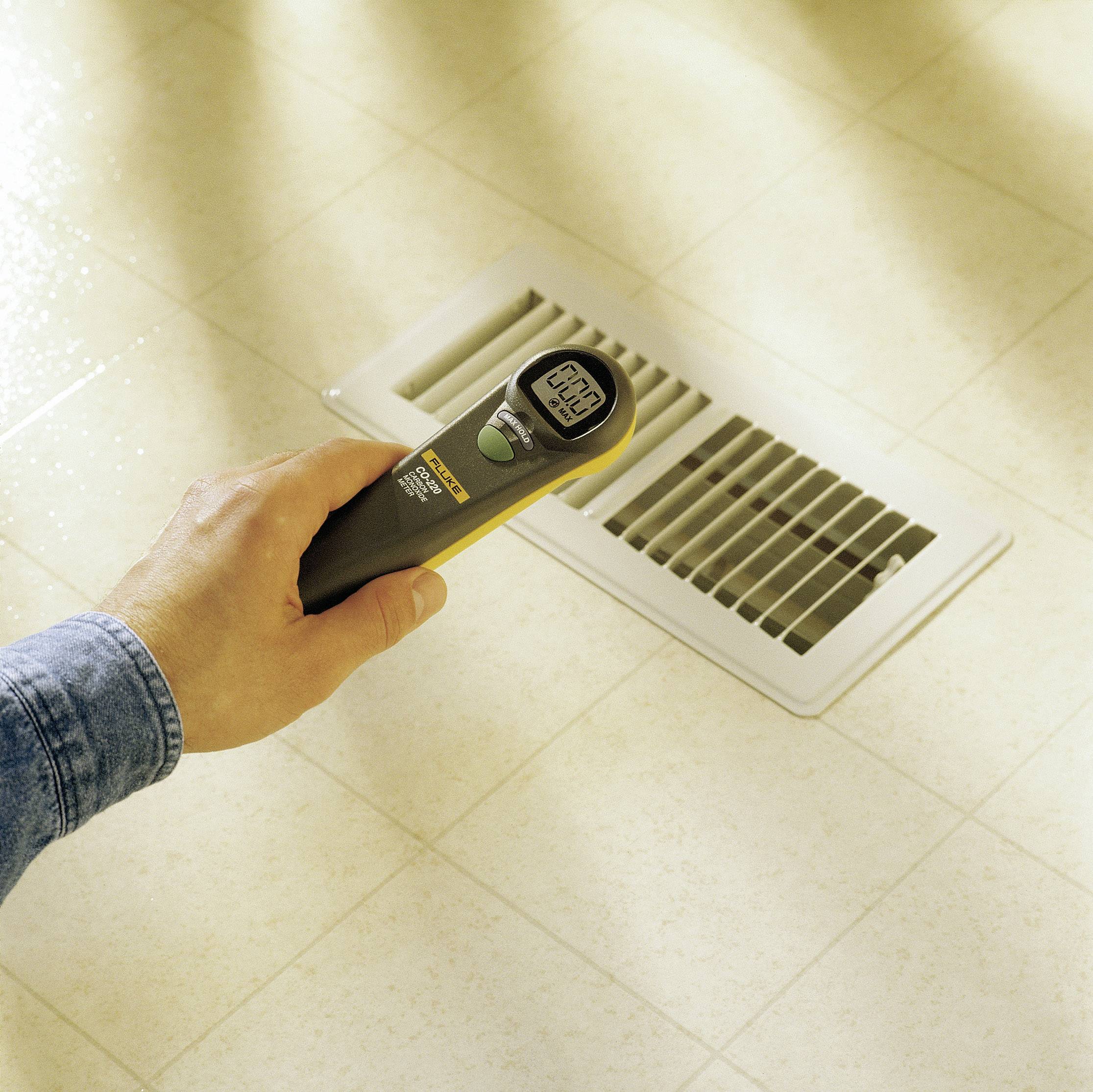 Una mano tiene un termometro a infrarossi sopra una fessura di ventilazione su una superficie piastrellata, il display mostra '72,3'.