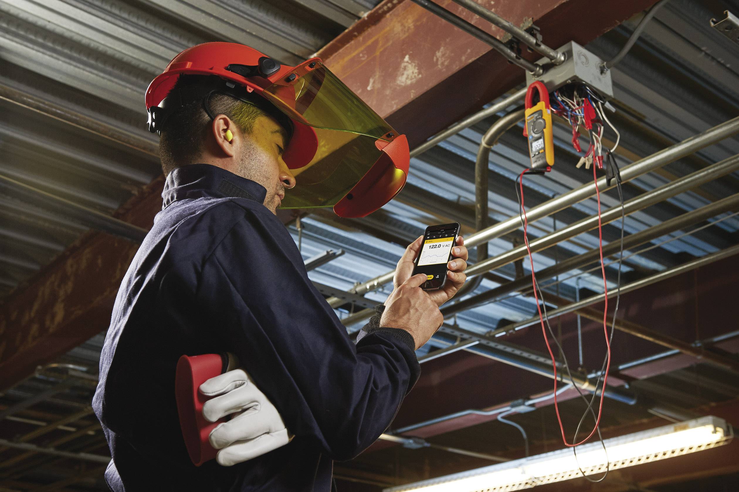 Un tecnico indossa dispositivi di protezione personale e utilizza uno strumento di misurazione per esaminare i cavi elettrici in un ambiente industriale.