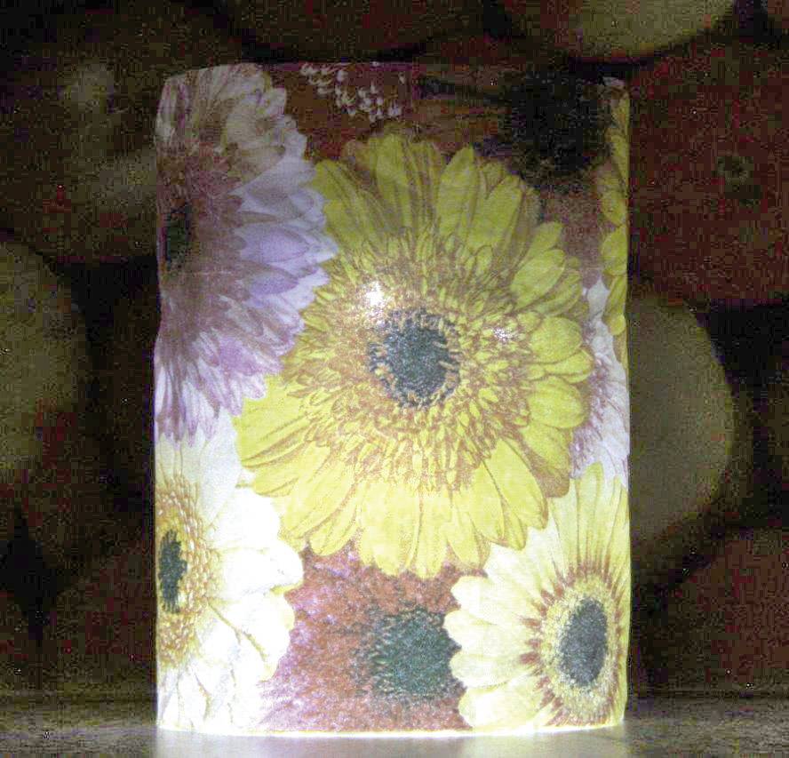 Una candela con un motivo floreale in tonalità di giallo, bianco e rosa è posizionata davanti a uno sfondo scuro.